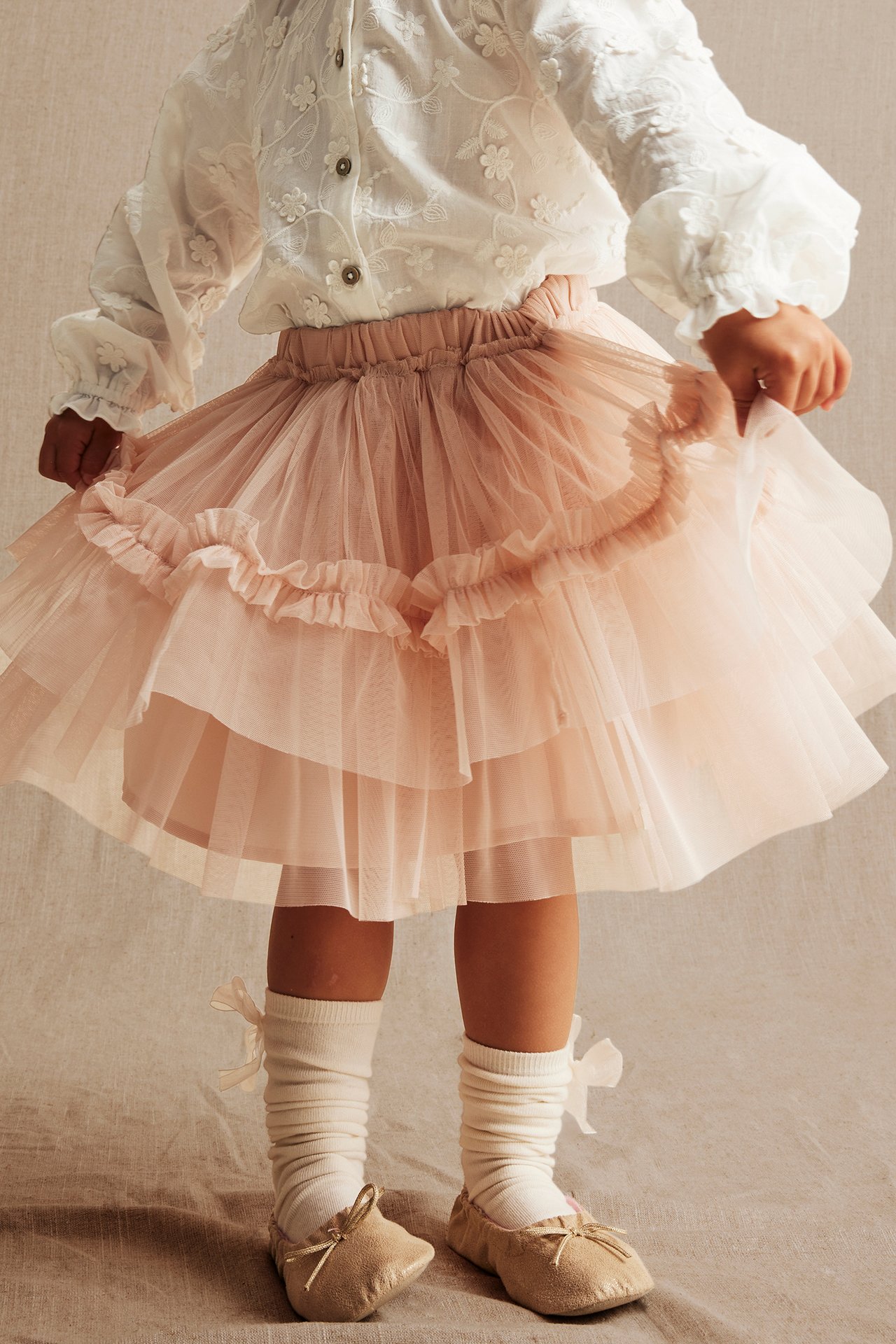 Front view of a kid in an offwhite embroidered blouse, pink layered tulle skirt, offwhite socks, and light brown shoes.