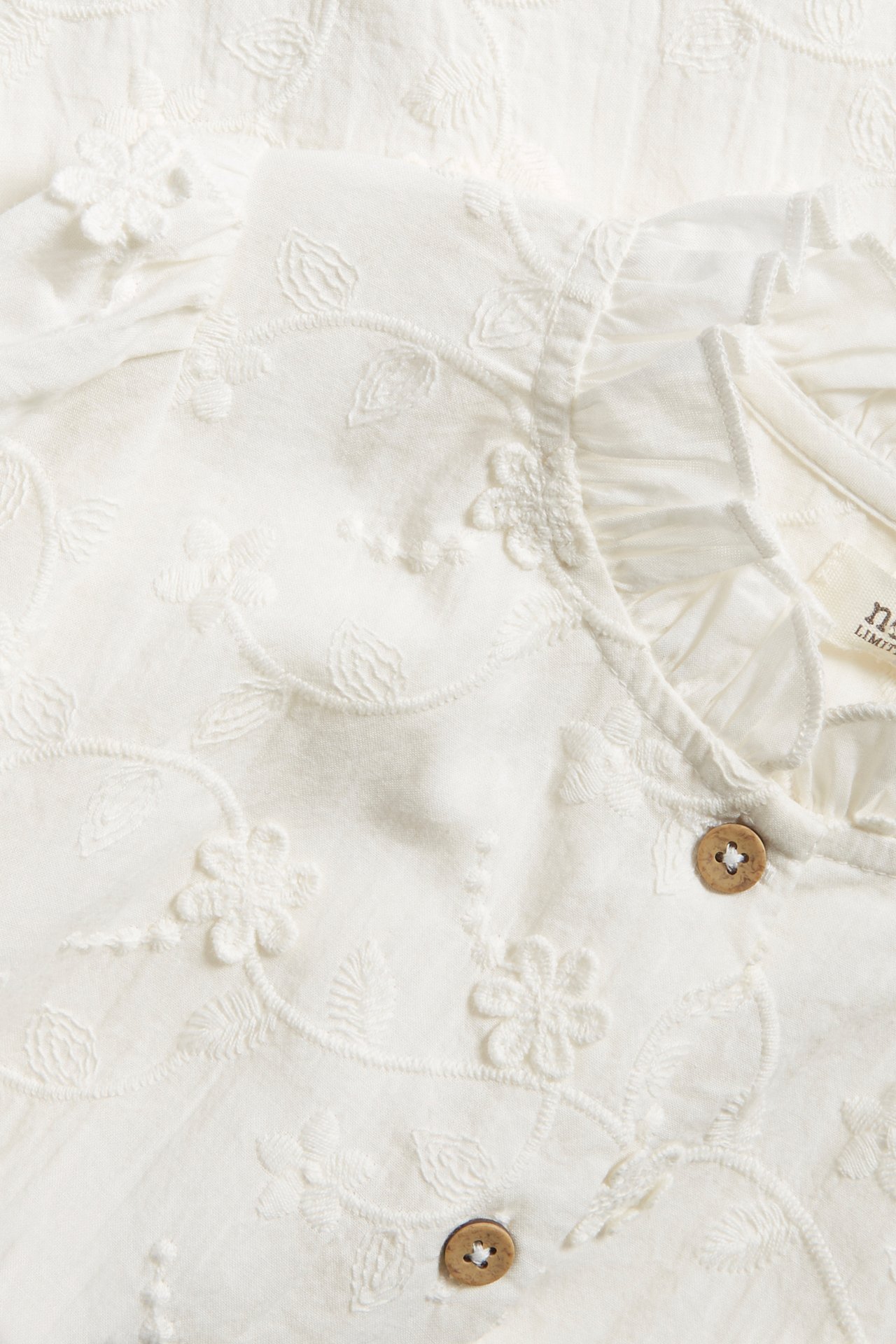 Close-up of an off-white organic cotton blouse with allover floral embroidery, a ruffled collar, and faux coconut buttons.