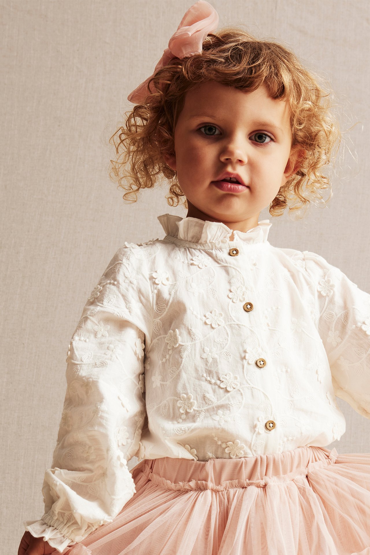 Front view: Kid in offwhite floral embroidered blouse with buttons, pink tulle skirt, and pink bow.