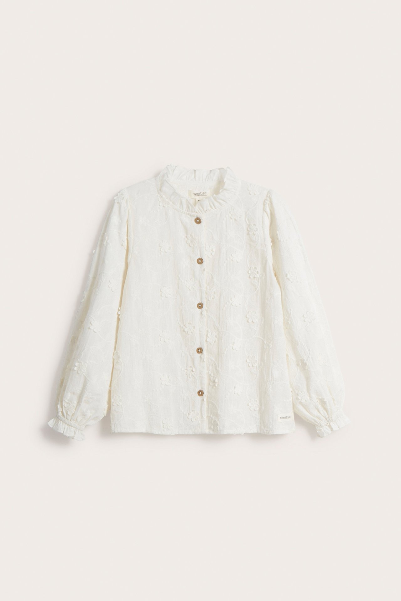 Front view of an off-white organic cotton blouse with allover 3D floral embroidery, ruffled collar, and brown buttons.