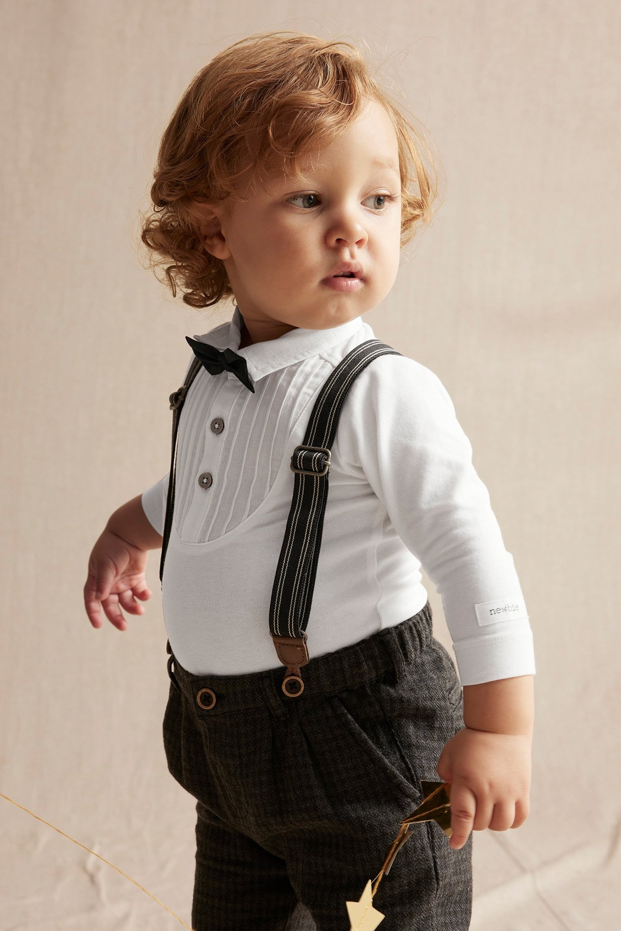 Baby boy in an off-white tuxedo bodysuit with a black bow tie, dark grey trousers, and black suspenders.