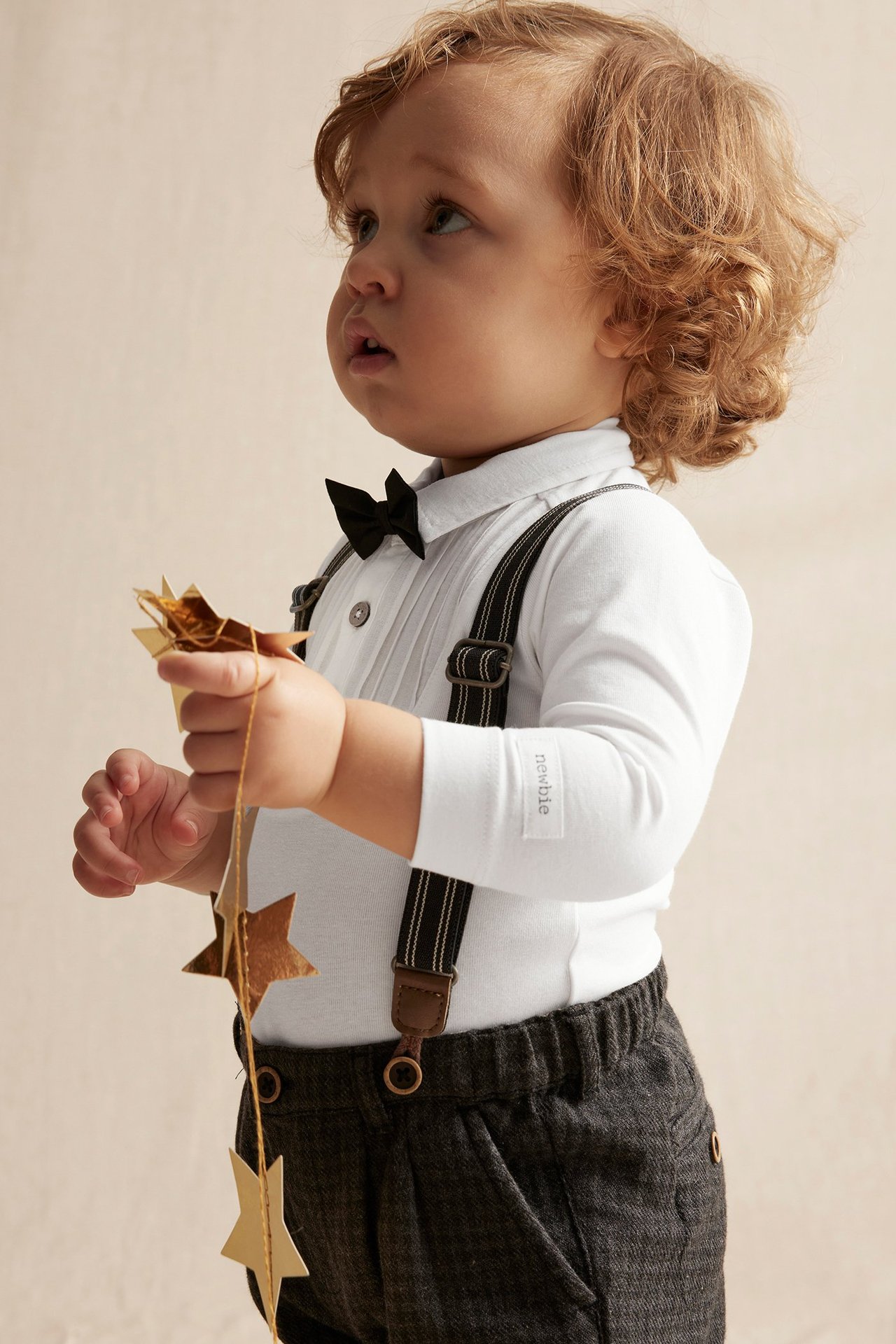 Side view: Baby in off-white long-sleeved bodysuit with black bow tie, grey trousers, suspenders, holding golden stars.