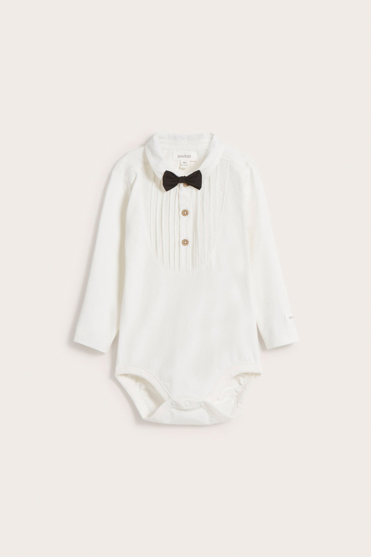 Front view: Off-white long-sleeved baby bodysuit with pleated front, snap closure, and black bow tie.