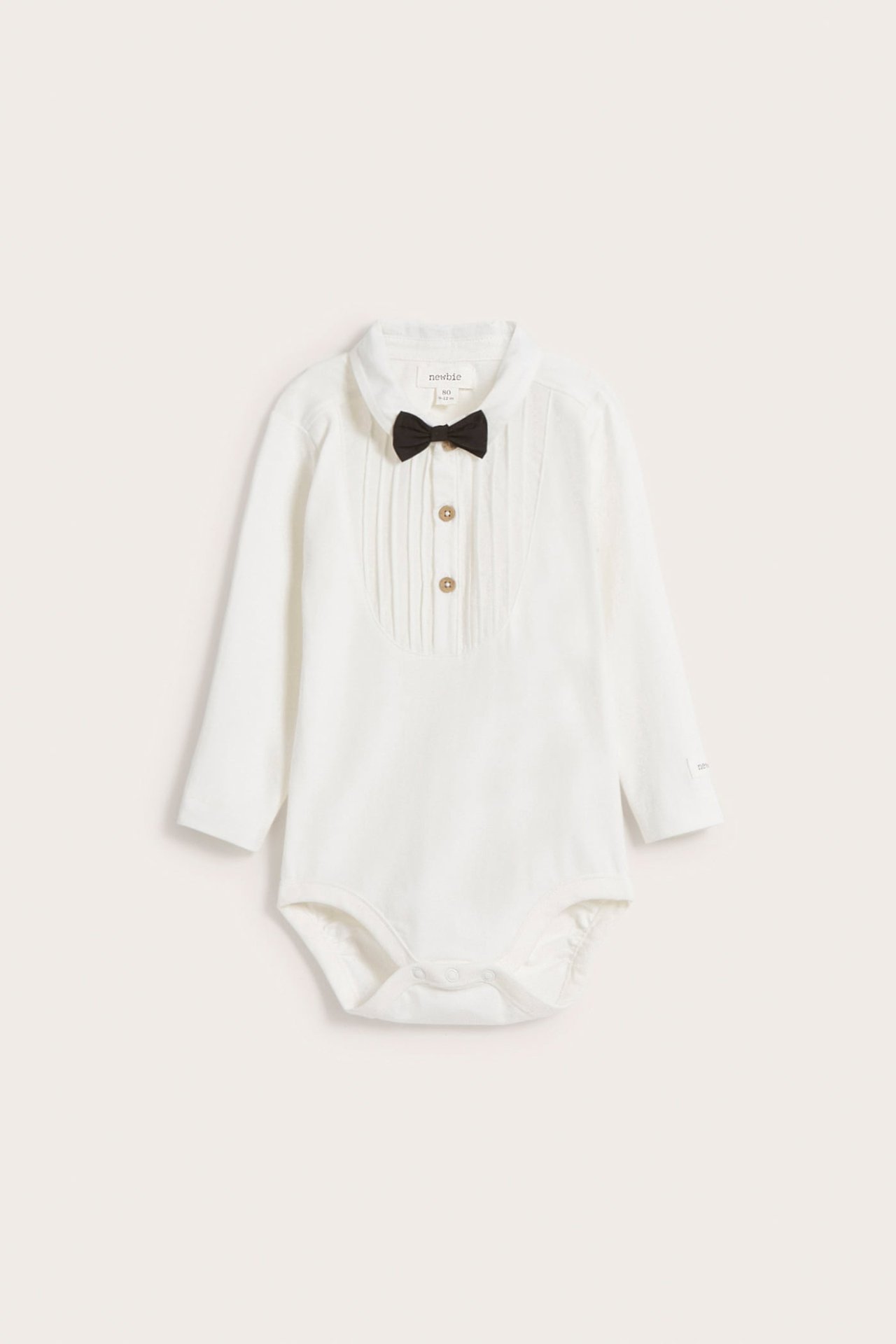 Vorderansicht: Cremefarbener langärmeliger Baby-Body mit plissierter Vorderseite, Druckknopfverschluss und schwarzer Fliege.