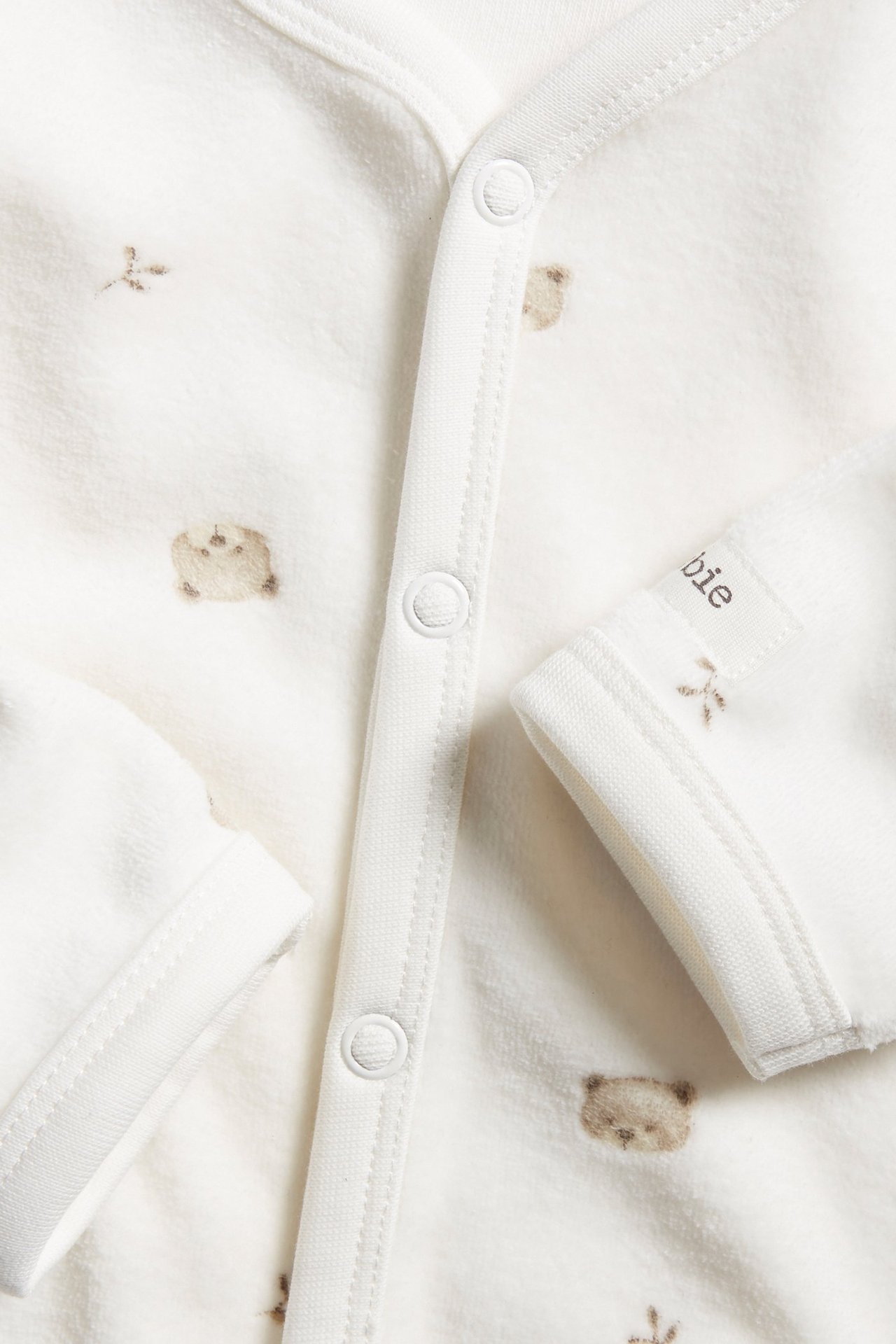 Nahaufnahme eines cremefarbenen Velours-Babystramplers mit Teddybärmuster und Druckknöpfen vorne.