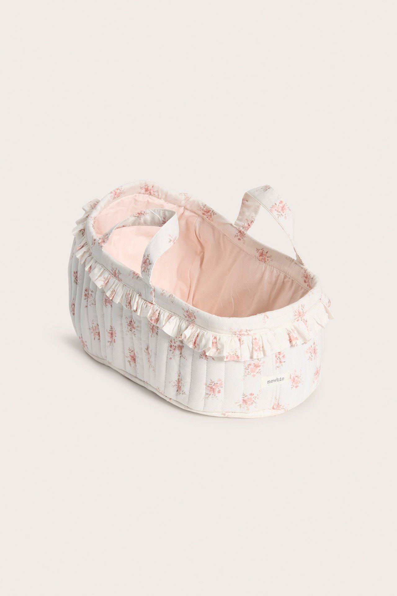 Schräge Vorderansicht: Off-weißer gesteppter Puppenkorb mit rosa Blumenmuster, hellrosa Futter und Rüschenrand.