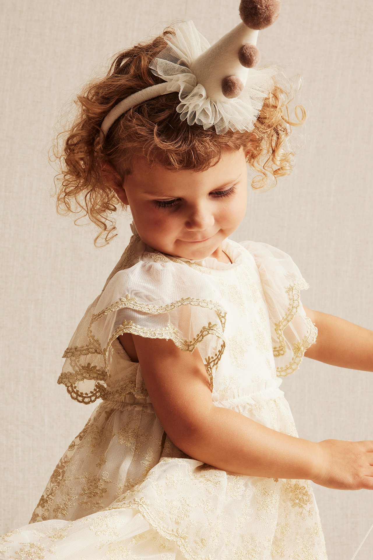 Nahaufnahme eines Kindes in einem offwhite bestickten Mesh-Kleid mit Golddetails und einem Partyhut-Haarreif.