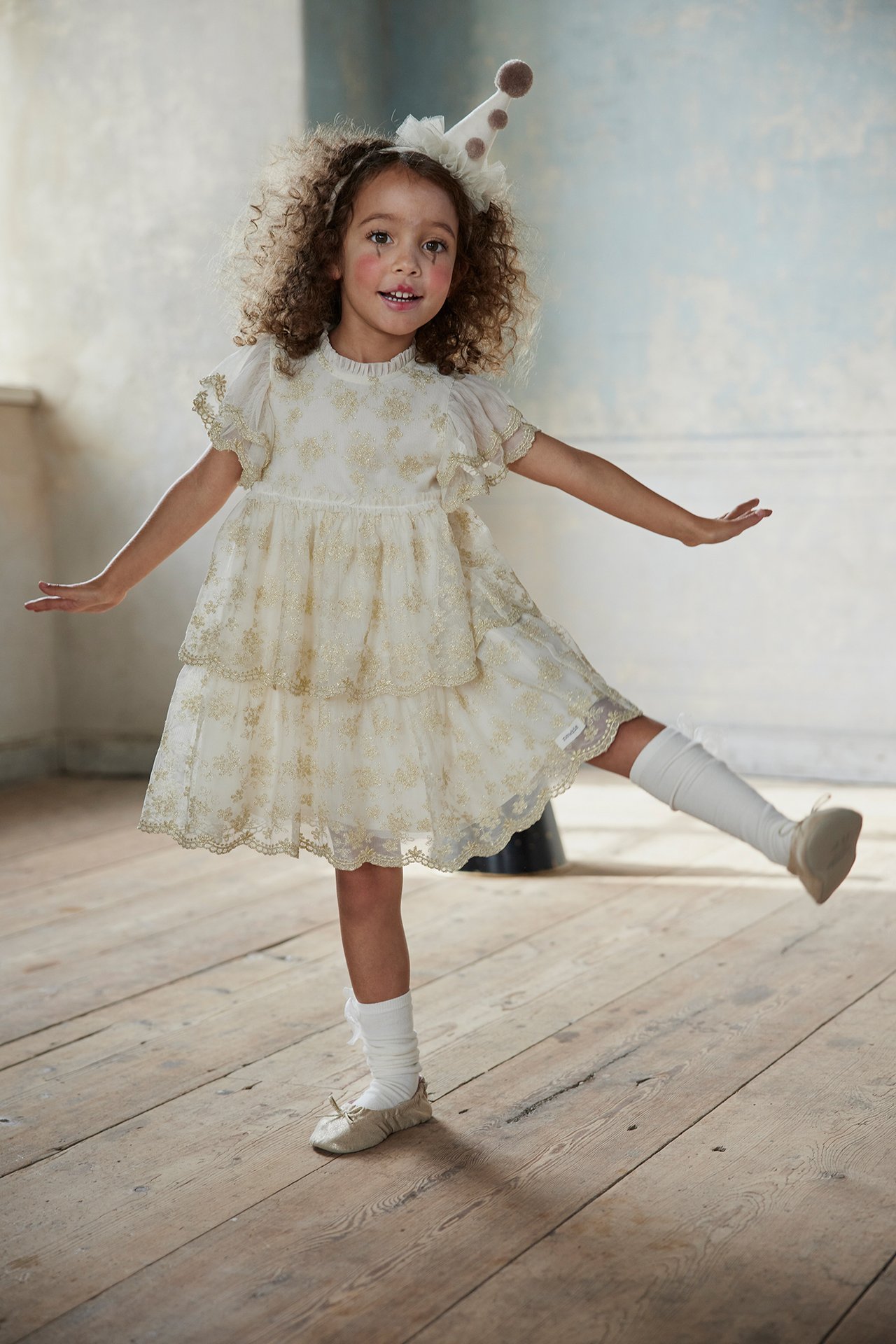 Vorderansicht Kind in cremefarbenem besticktem Mesh-Kleid mit Golddetails, weißen Socken und beigen Schuhen.