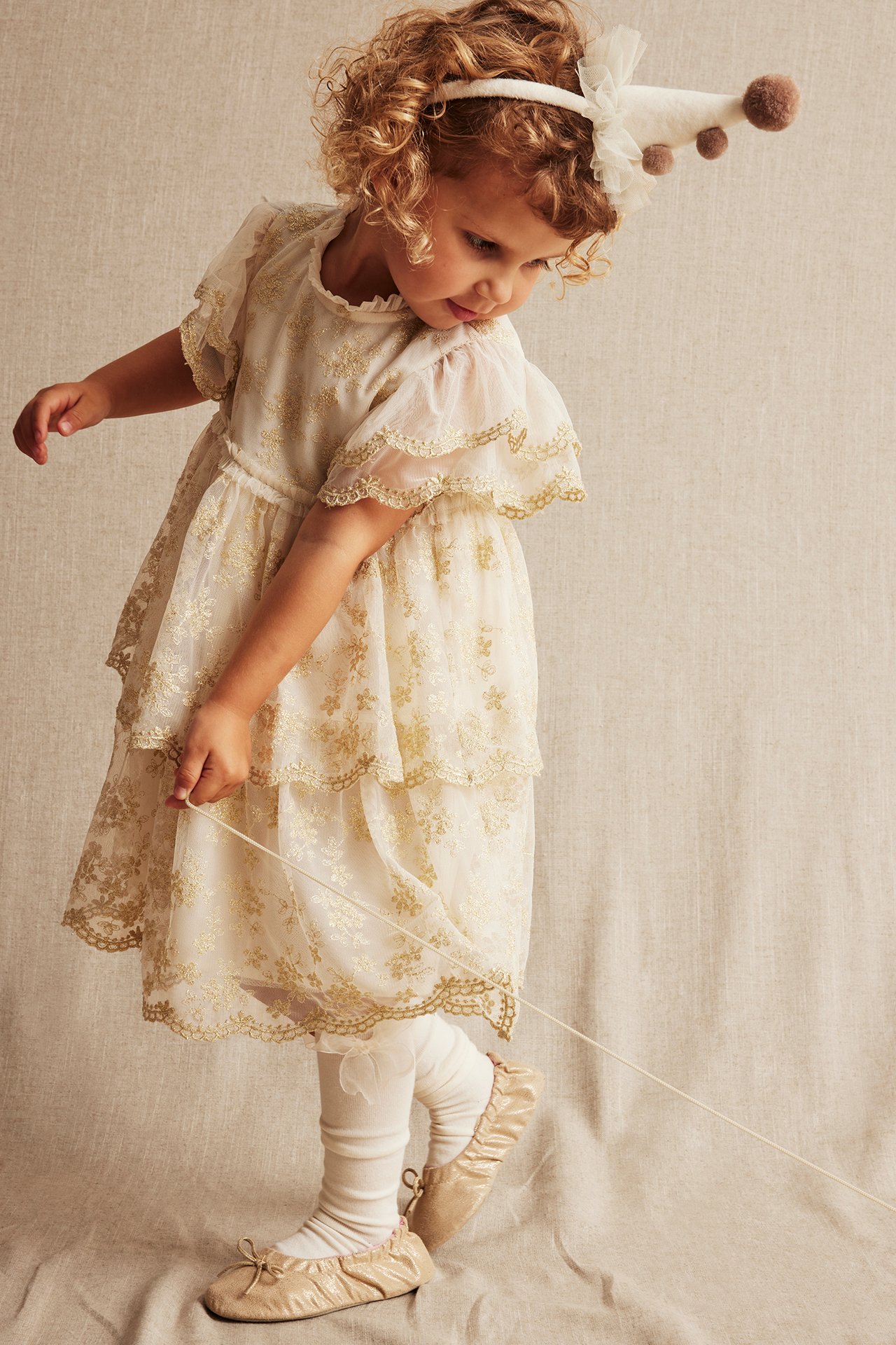 Vorderansicht eines Kindes in einem cremefarbenen bestickten Mesh-Kleid, weißen Strumpfhosen, beigen Ballerinas und einem Partyhut.