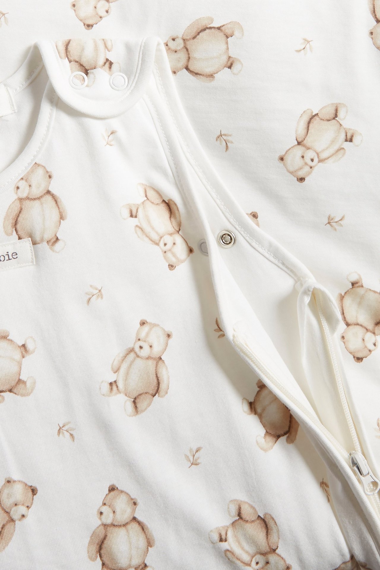 Nahaufnahme eines cremefarbenen Babyschlafsacks mit braunem Teddybärenmuster, Druckknöpfen und Zwei-Wege-Reißverschluss.