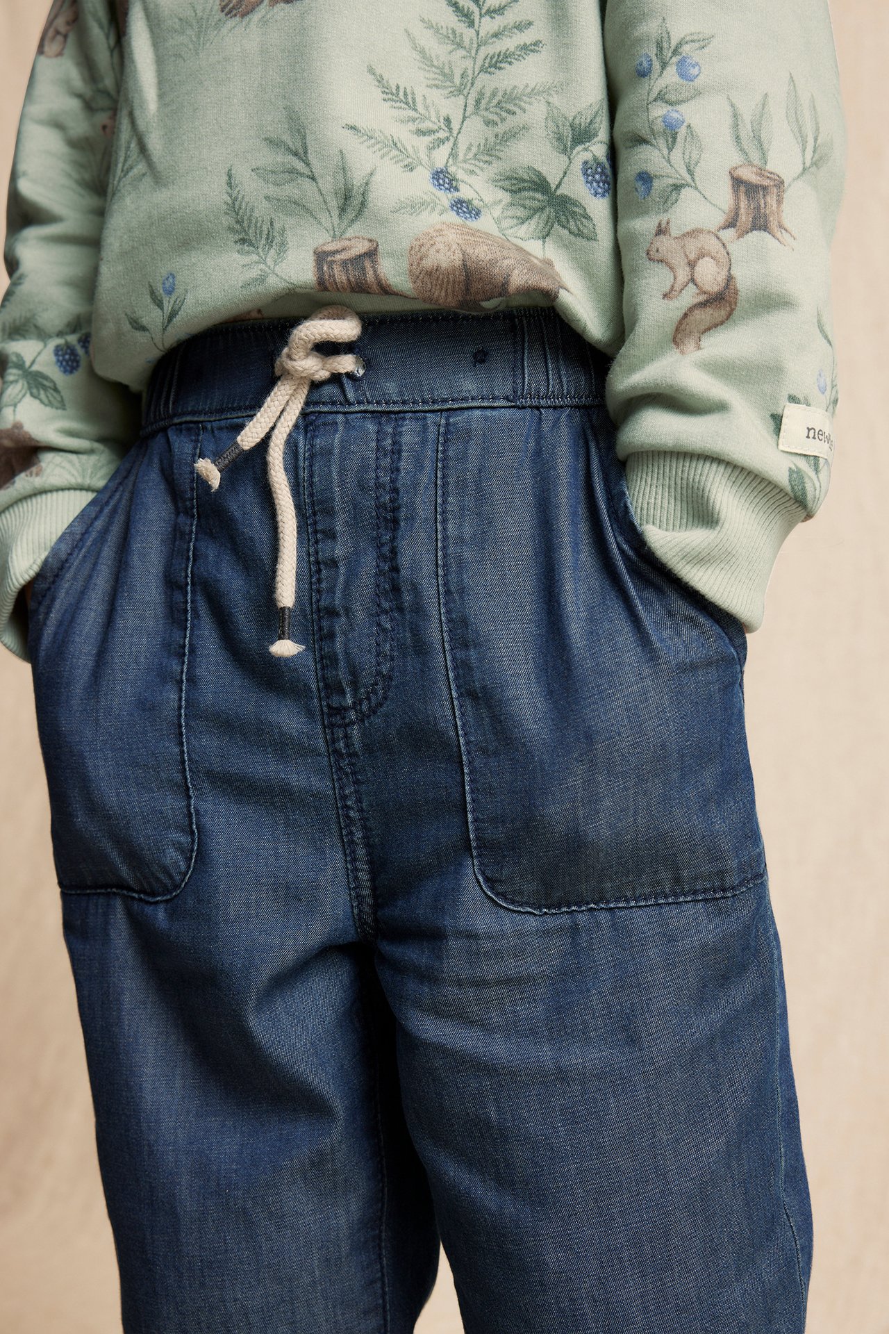 Close-up of a kid wearing blue cotton jeans with drawstring waist and patch pockets, and a green patterned sweatshirt.