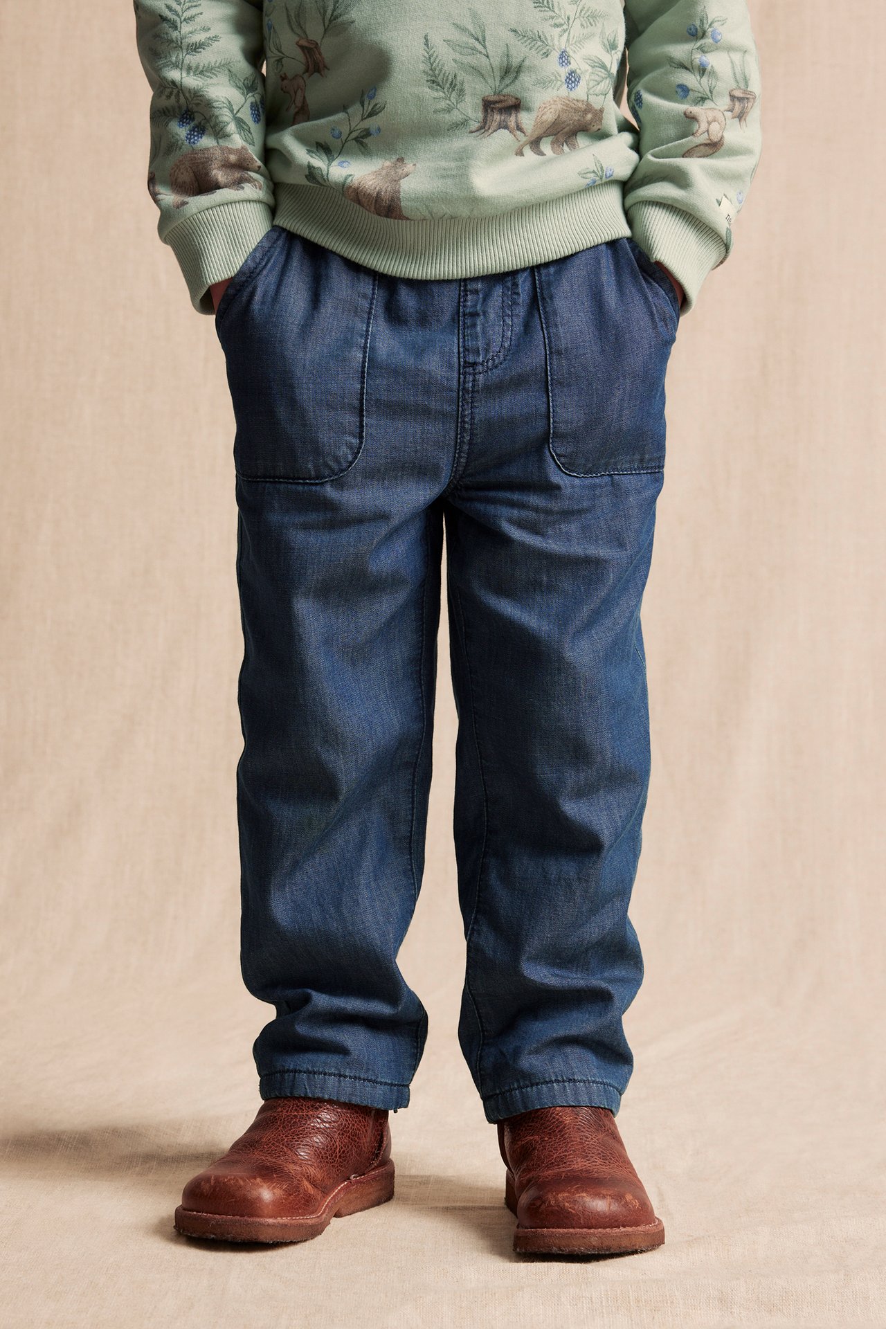 Front view of a kid wearing blue cotton jeans with front pockets, a patterned sweatshirt, and brown boots.