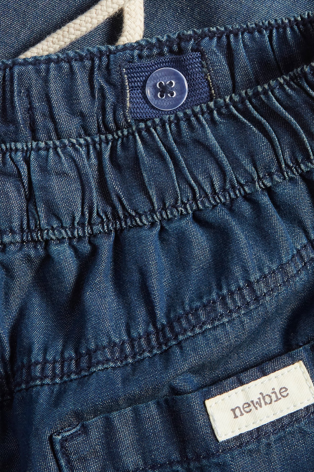 Close-up of blue cotton jeans with an elasticated drawstring waist, a button, and a Newbie label.