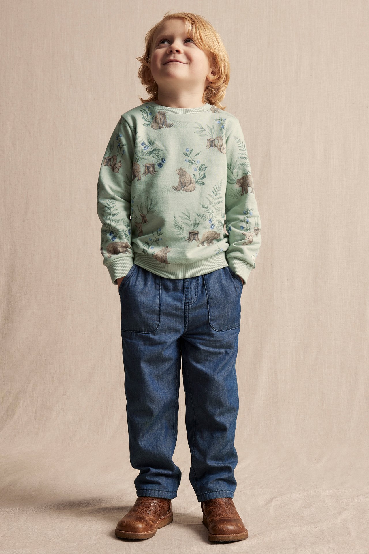 Front view of a kid in blue jeans with front pockets, a light green patterned sweatshirt, and brown boots.