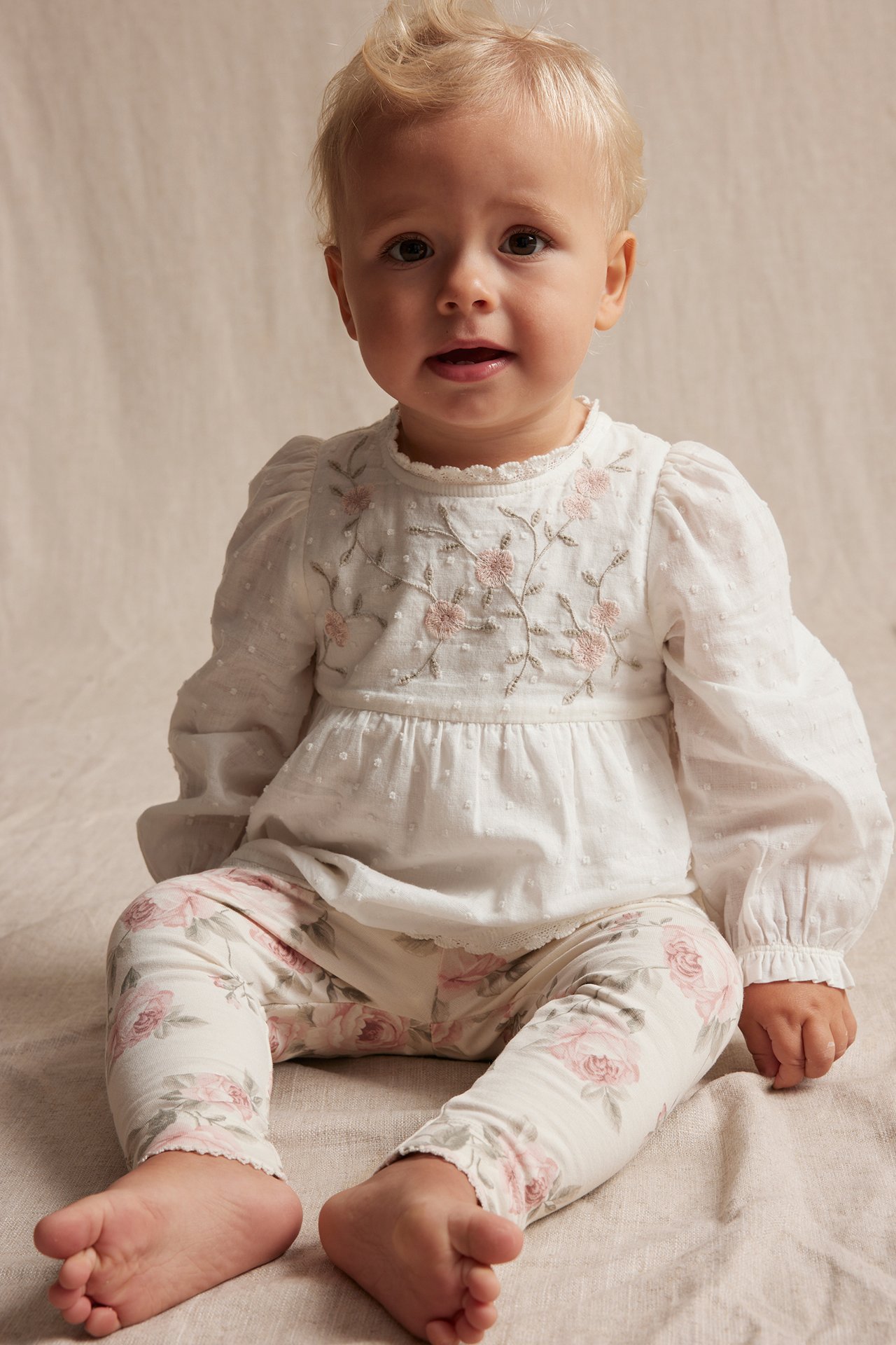 Front view of a baby wearing an off-white woven bodysuit with pink floral embroidery and white floral print leggings.