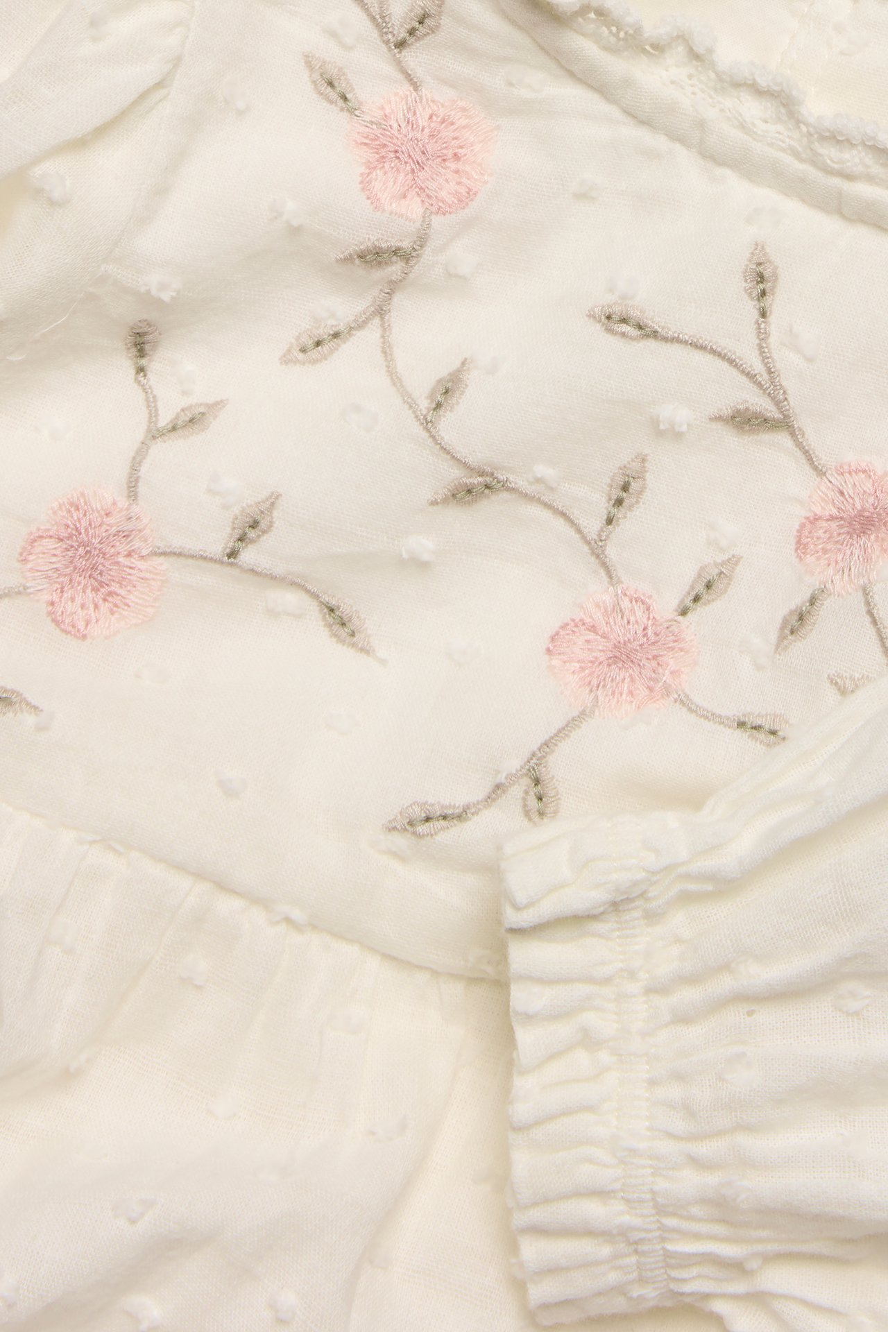 Close-up of an off-white woven baby bodysuit with textured dots, pink floral embroidery, and a lace-trimmed neckline.