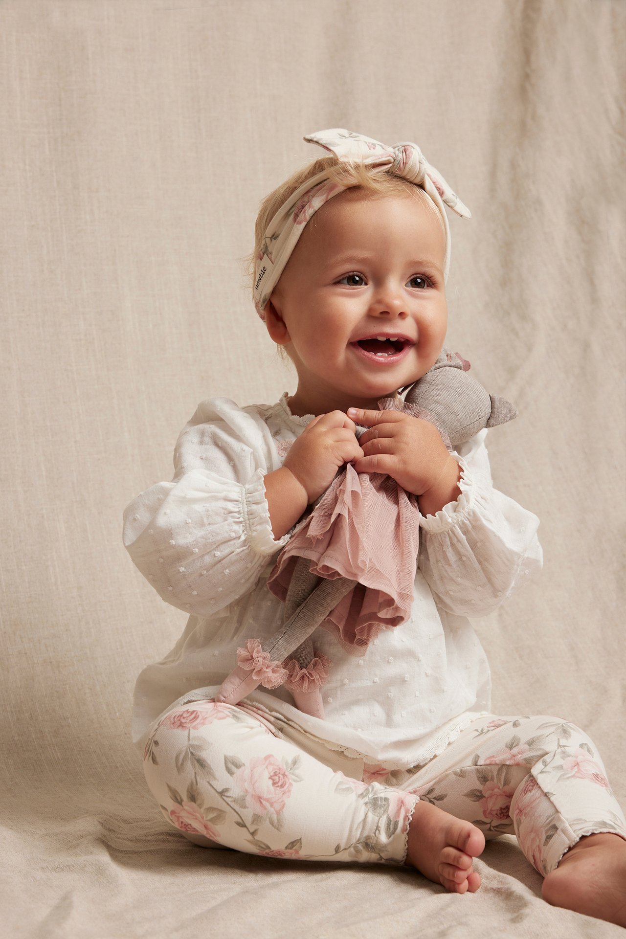 Front view of a smiling baby wearing an off-white woven body, floral leggings, and a matching headband, holding a doll.