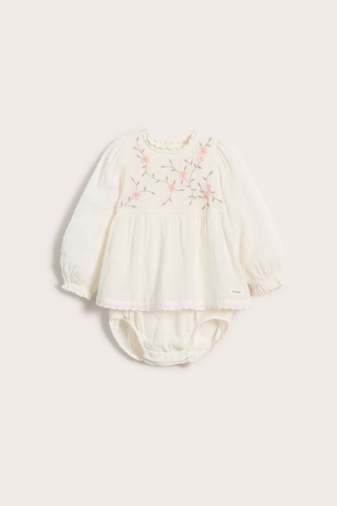 Front view of an offwhite woven baby bodysuit with texture dots, pink floral embroidery, puff sleeves, and lace details.