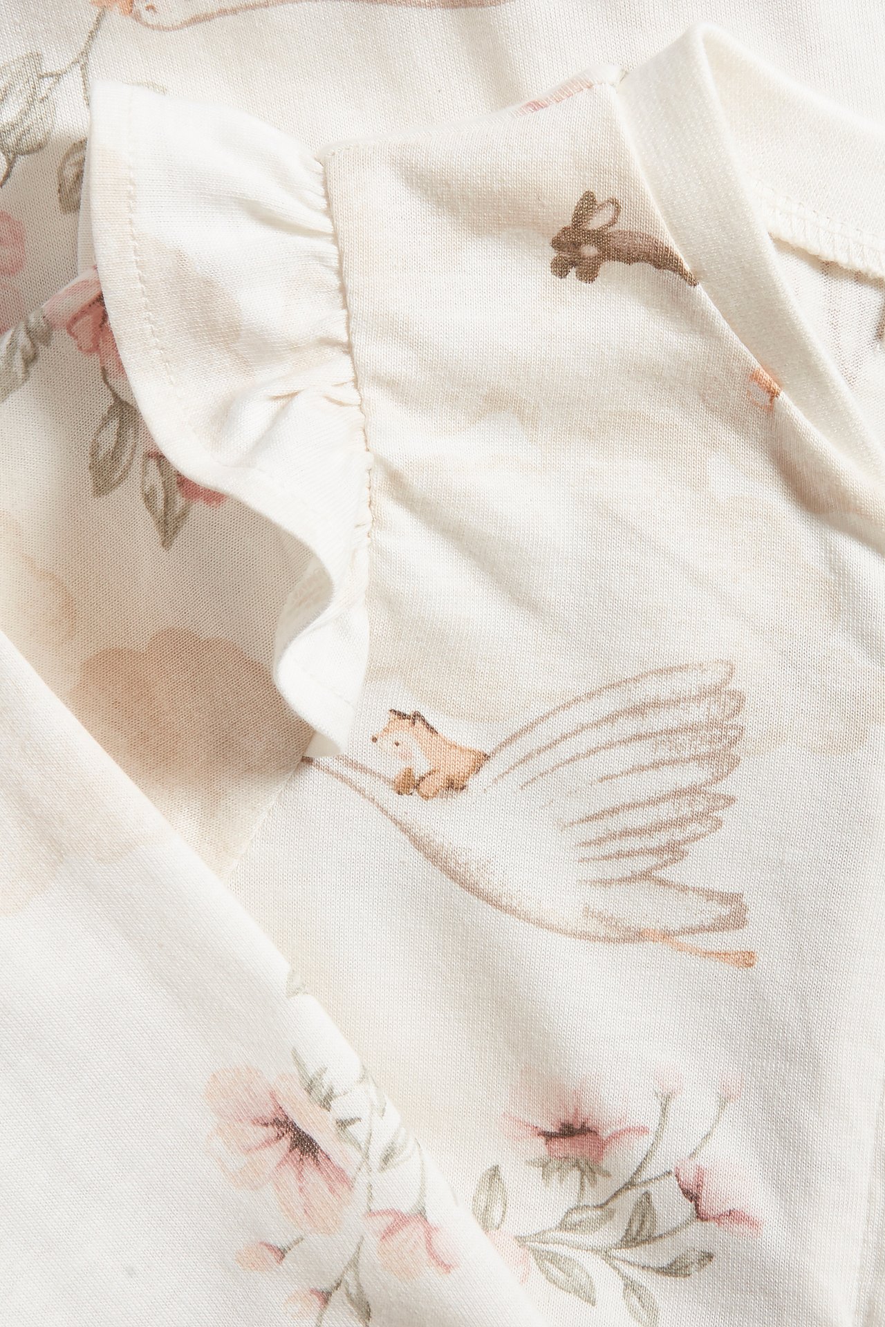 Nahaufnahme eines cremefarbenen Baby-Baumwollpyjamas mit Rüschen und niedlichem Tier- und Blumenmuster.