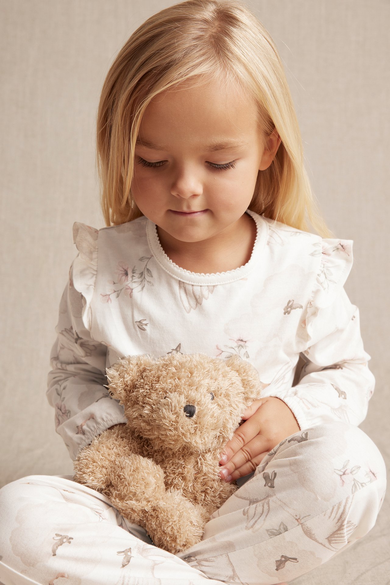 Front view of a kid in offwhite cotton pyjamas with ruffles and animal print, holding a teddy bear.