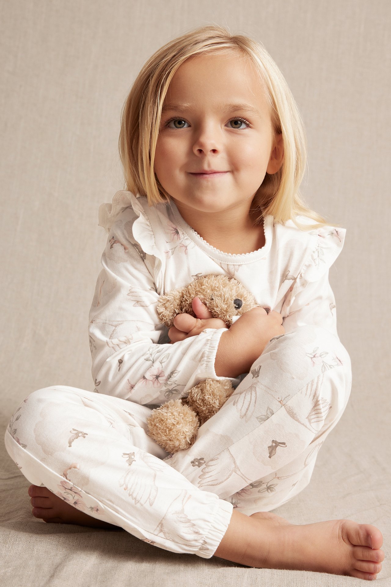 Front view of a kid in offwhite cotton pyjamas with ruffles and nature print, holding a teddy bear.