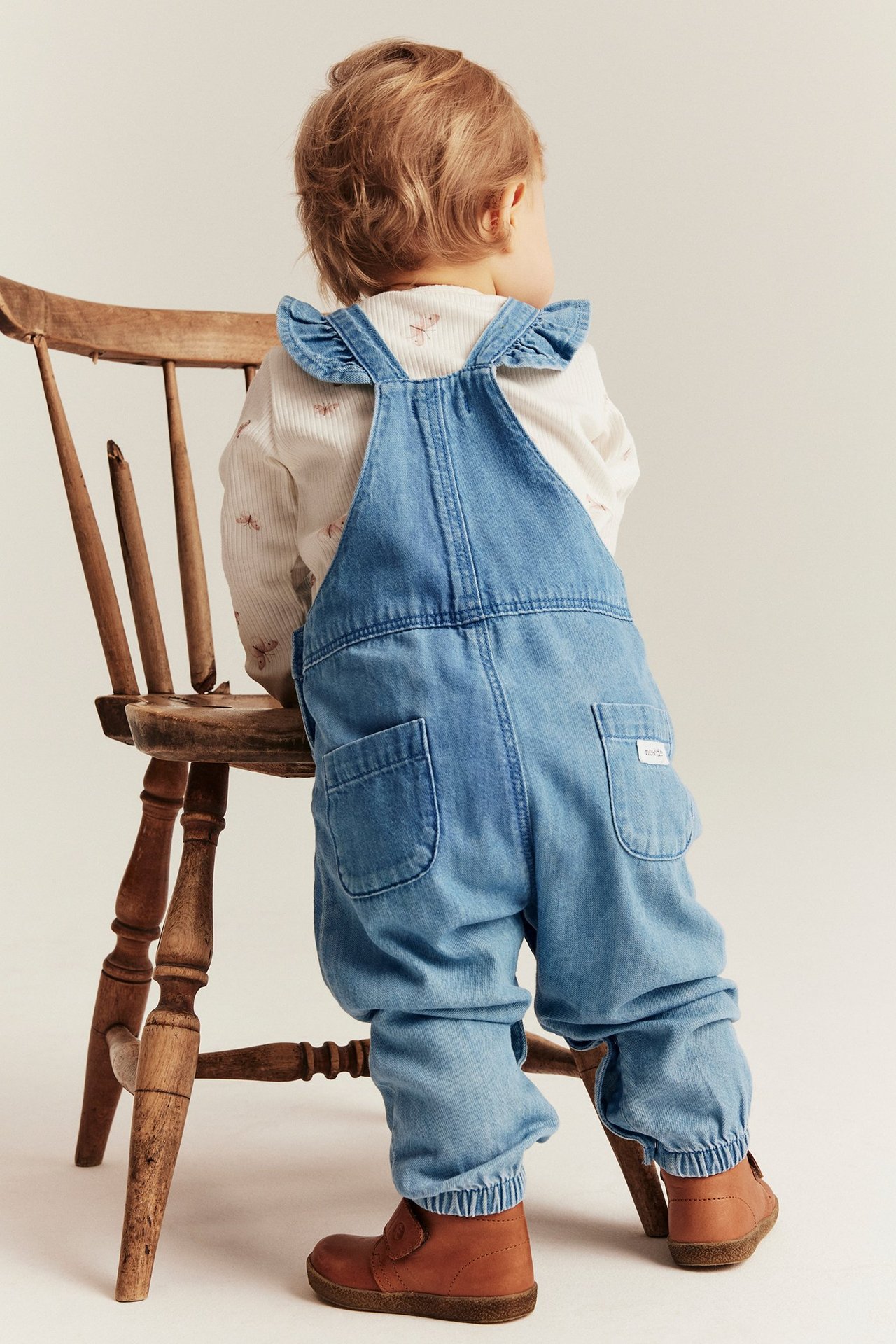 Rückenansicht eines Babys in hellblauen Denim-Latzhosen mit Rüschen-Trägern, dazu weißes gemustertes Oberteil und braune Schuhe.