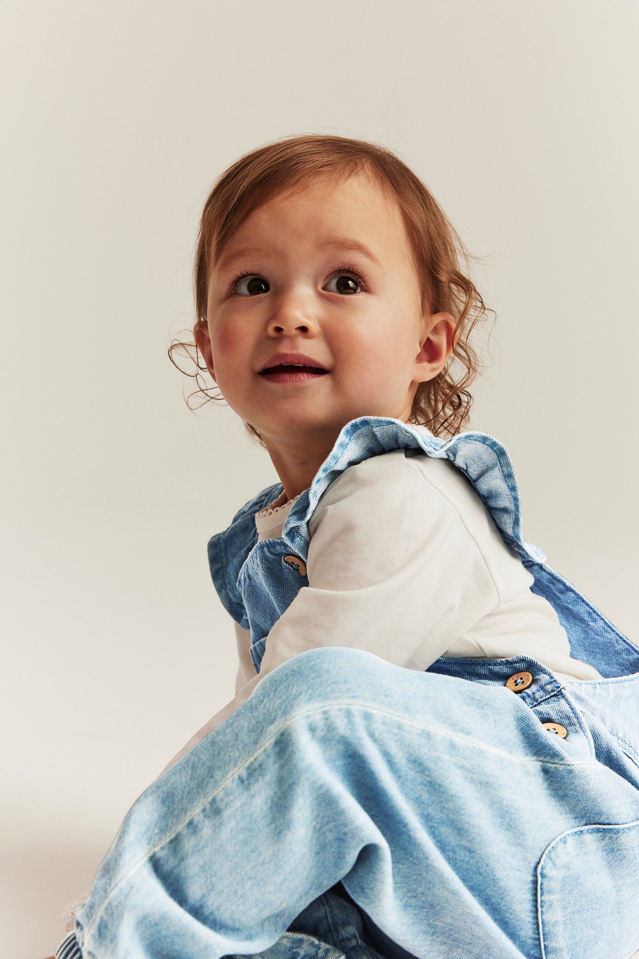 Close-up of a baby wearing light denim dungarees with ruffled straps over a white long-sleeved top.