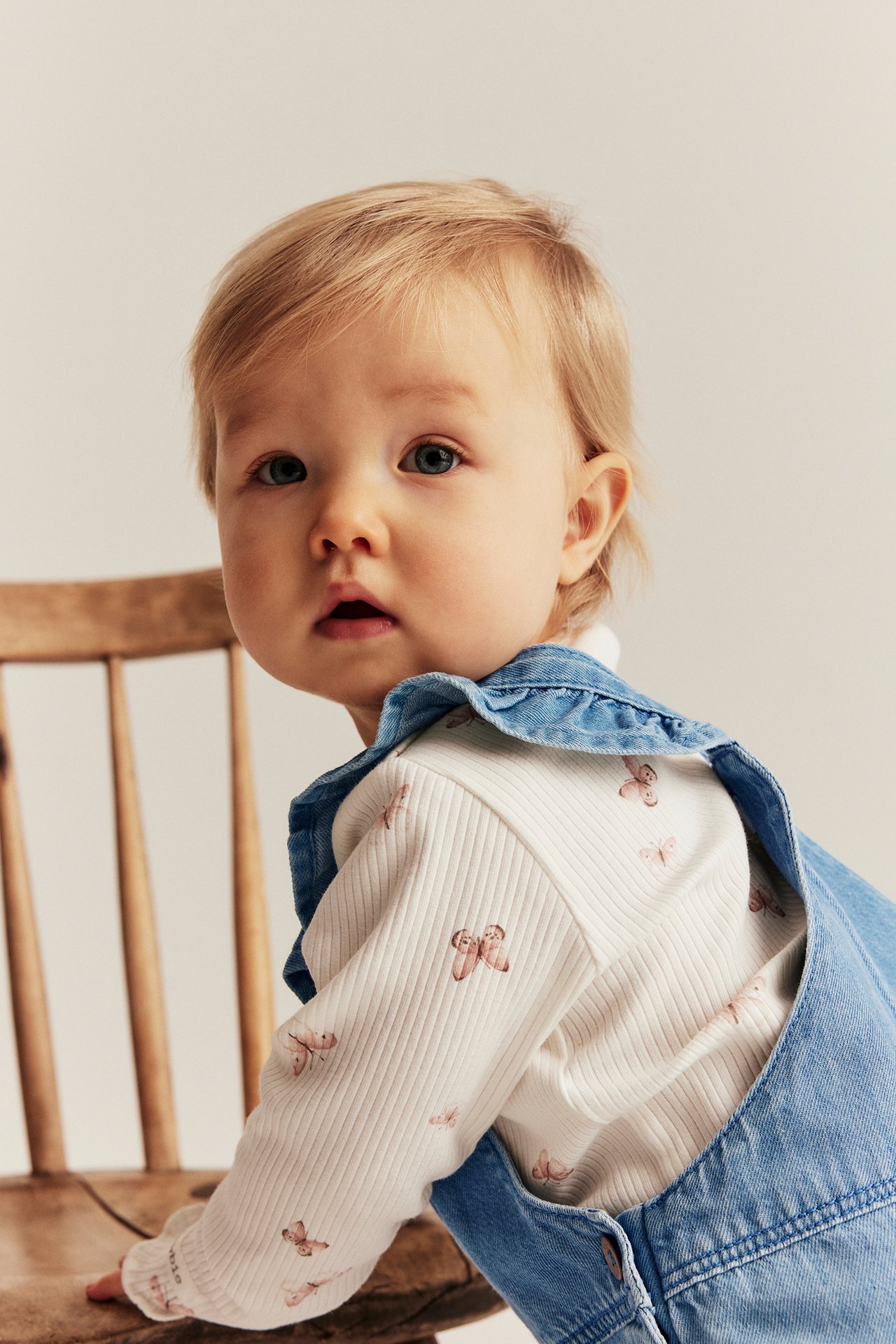 Close-up of a baby wearing light blue denim overalls with ruffled straps over a white long-sleeve top with butterfly print.