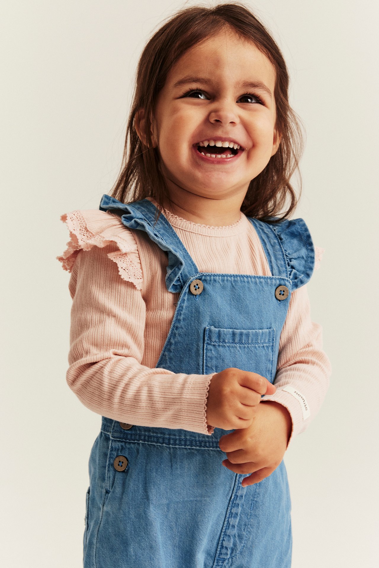 Vorderansicht eines Kindes in hellblauer Jeanslatzhose mit Holzknöpfen über einem rosa Langarmshirt.
