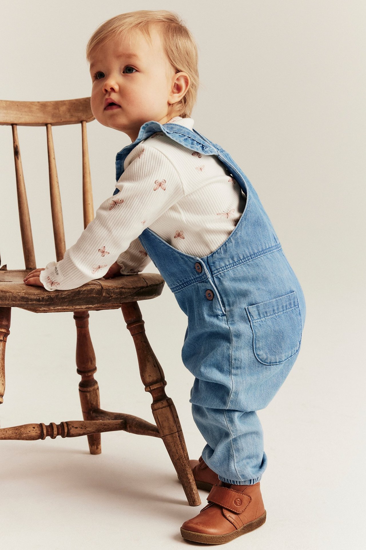 Side view: baby in light blue ruffled denim overalls over a white butterfly print top and brown boots.