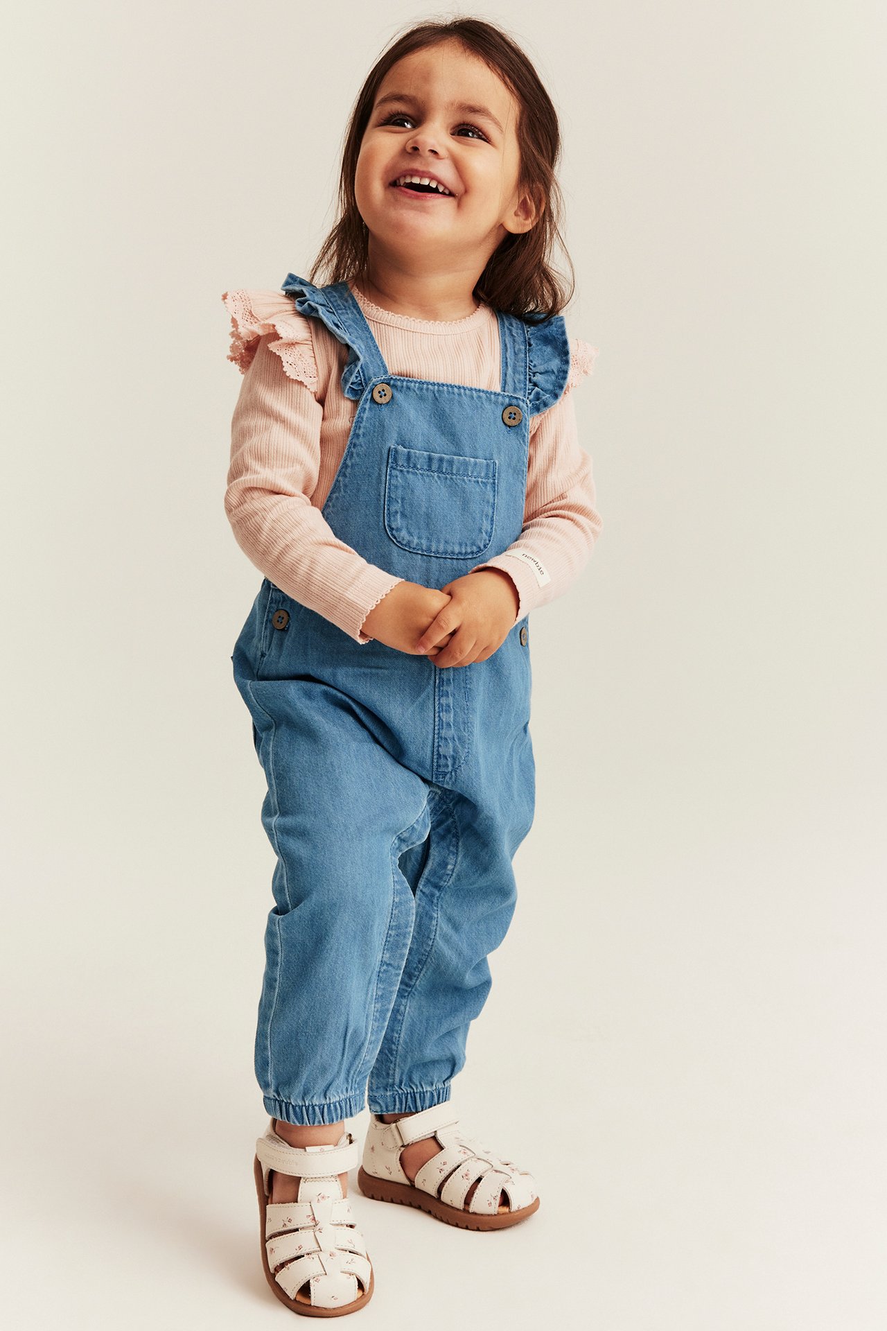 Vorderansicht: Kind in blauen Jeanslatzhosen mit Rüschen, rosa Rippentop und weißen gemusterten Sandalen.