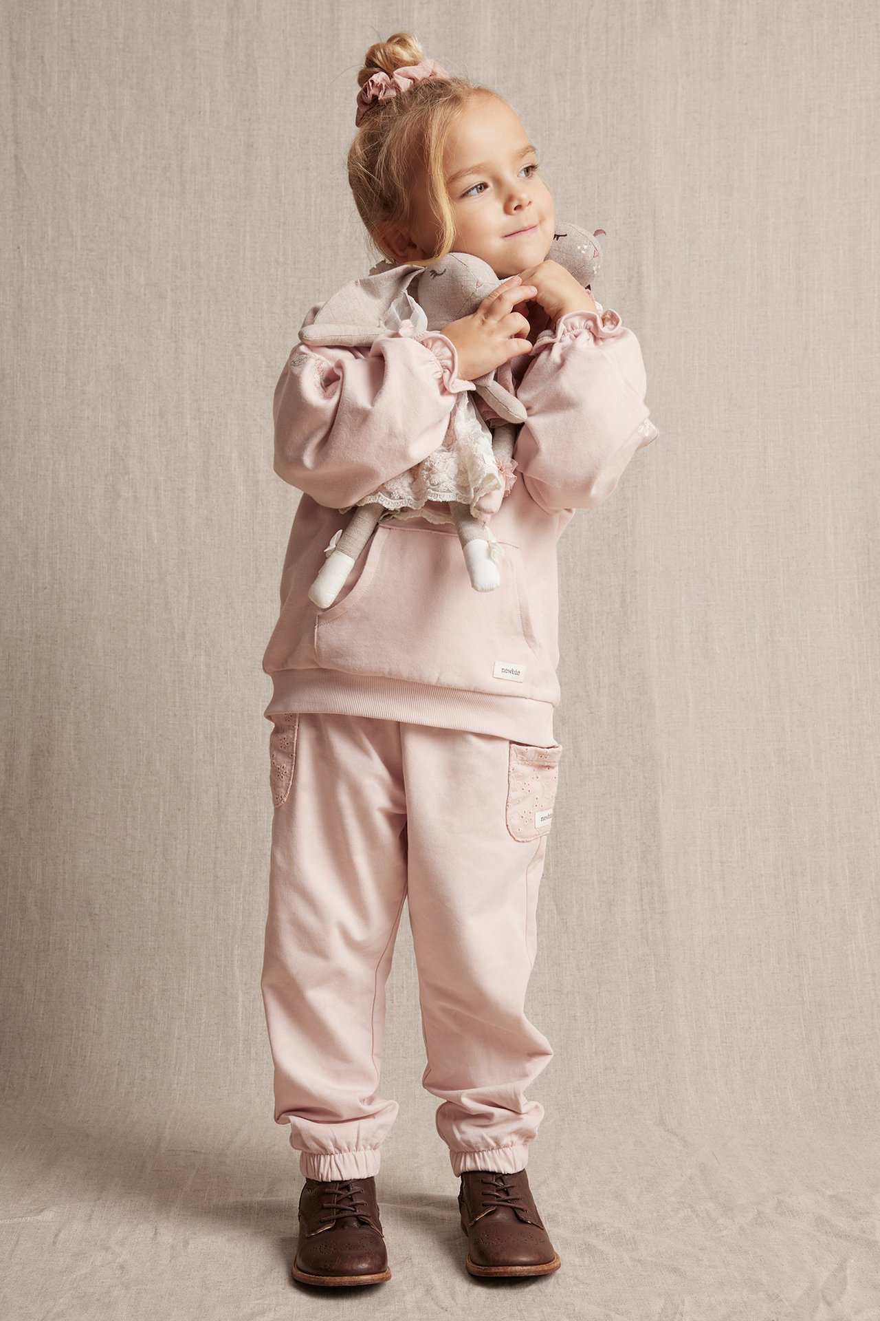 Front view: Kid in light pink cotton jogging pants with embroidered pockets, matching hoodie, and brown shoes, holding a doll.