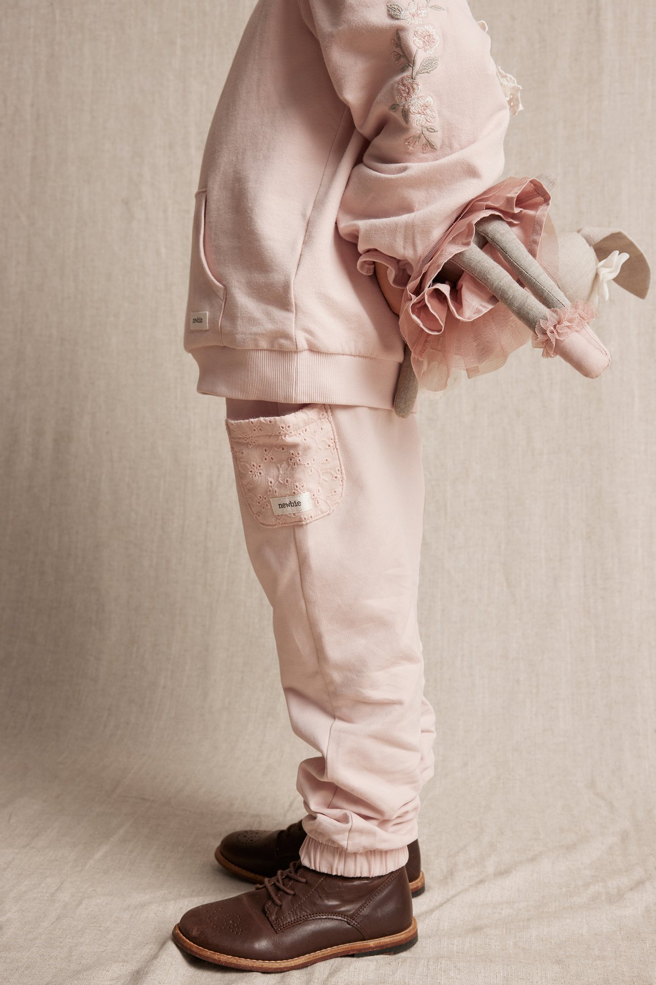 Side view of a kid in light pink jogging pants with embroidered pockets, pink hoodie, brown shoes, holding a doll.