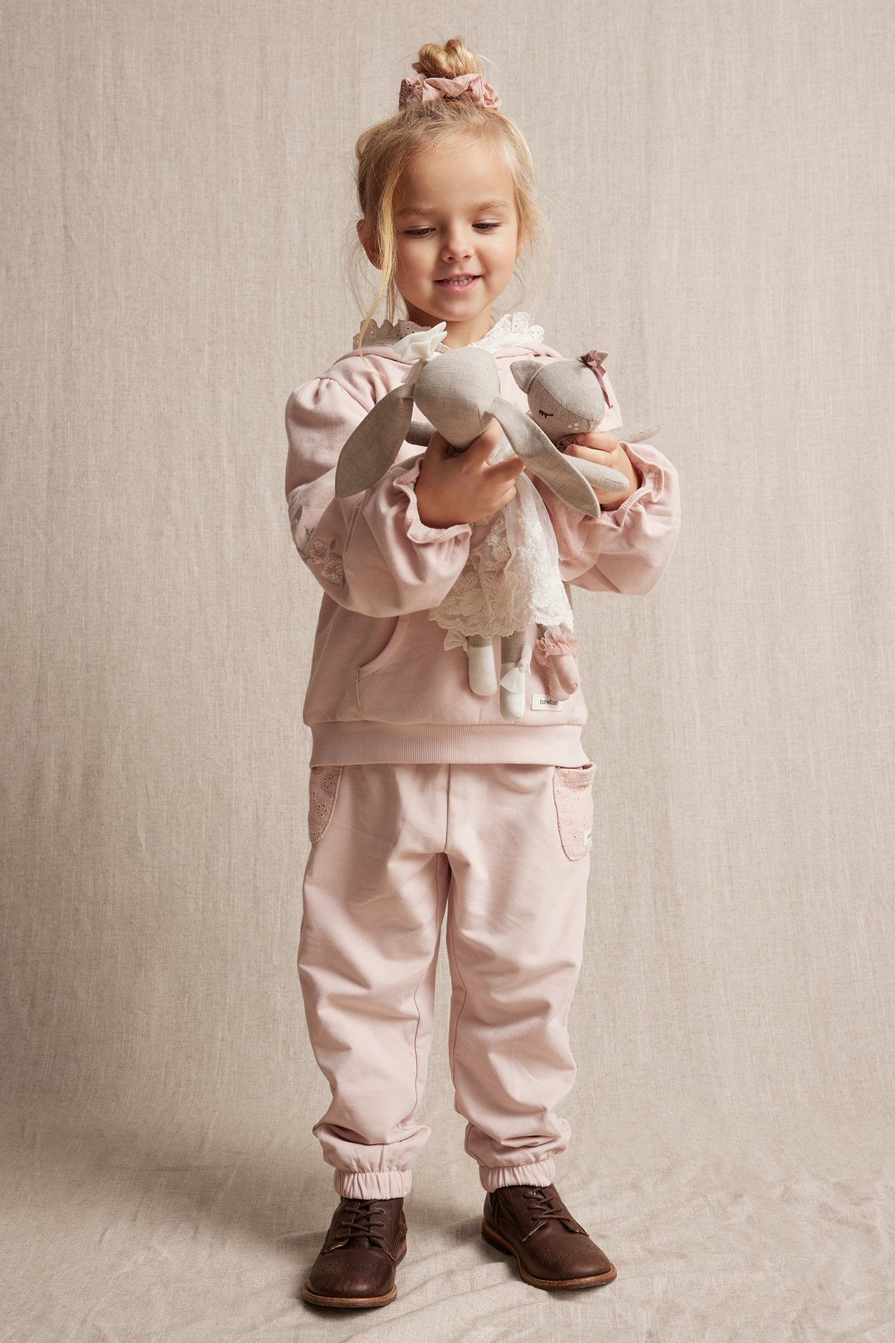 Front view of a kid in light pink cotton jogging pants with embroidered pockets, matching hoodie, and brown shoes.