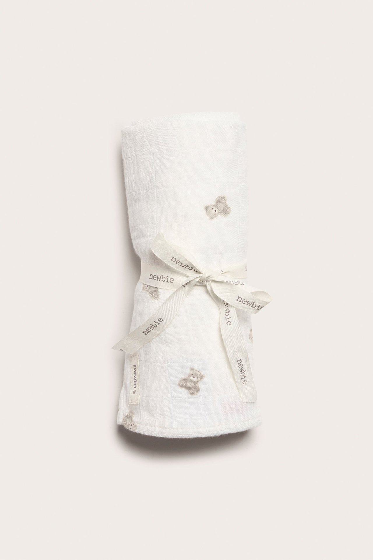 Front view of a rolled off-white muslin blanket with teddy bear pattern, tied with a "newbie" ribbon.