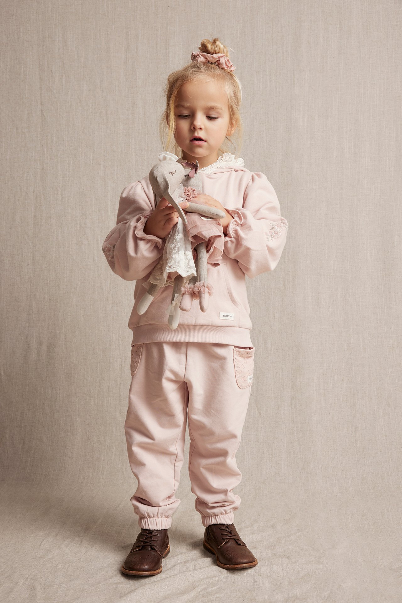 Front view of kid in light pink cotton hoodie with embroidery, lace hood, matching sweatpants, holding doll.
