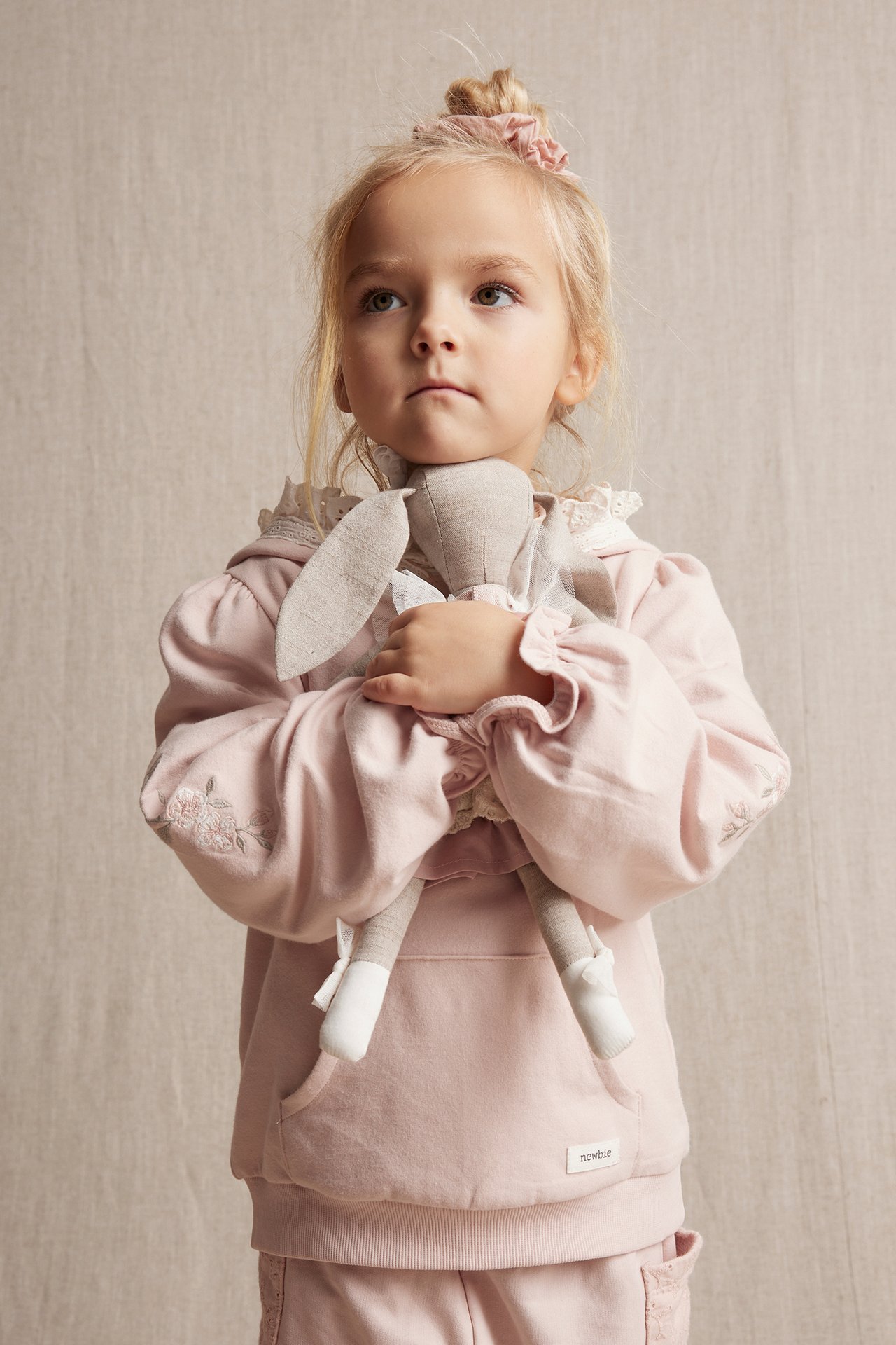 Front view: Kid in a light pink cotton hoodie with embroidered flowers, lace trim, holding a doll.