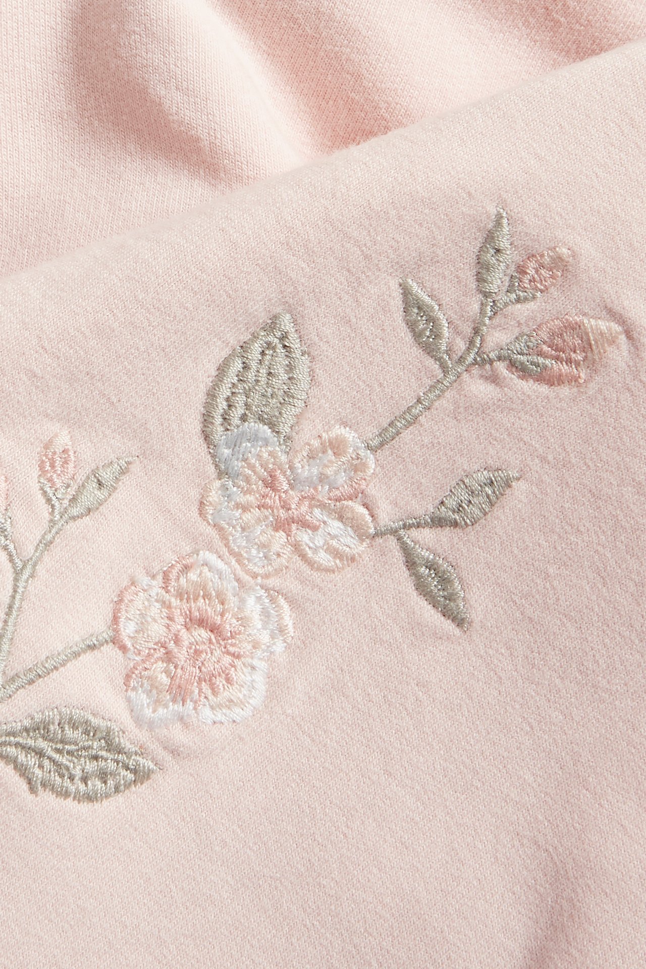 Close-up of a light pink cotton hoodie with delicate embroidered light pink and white flowers and grey leaves.