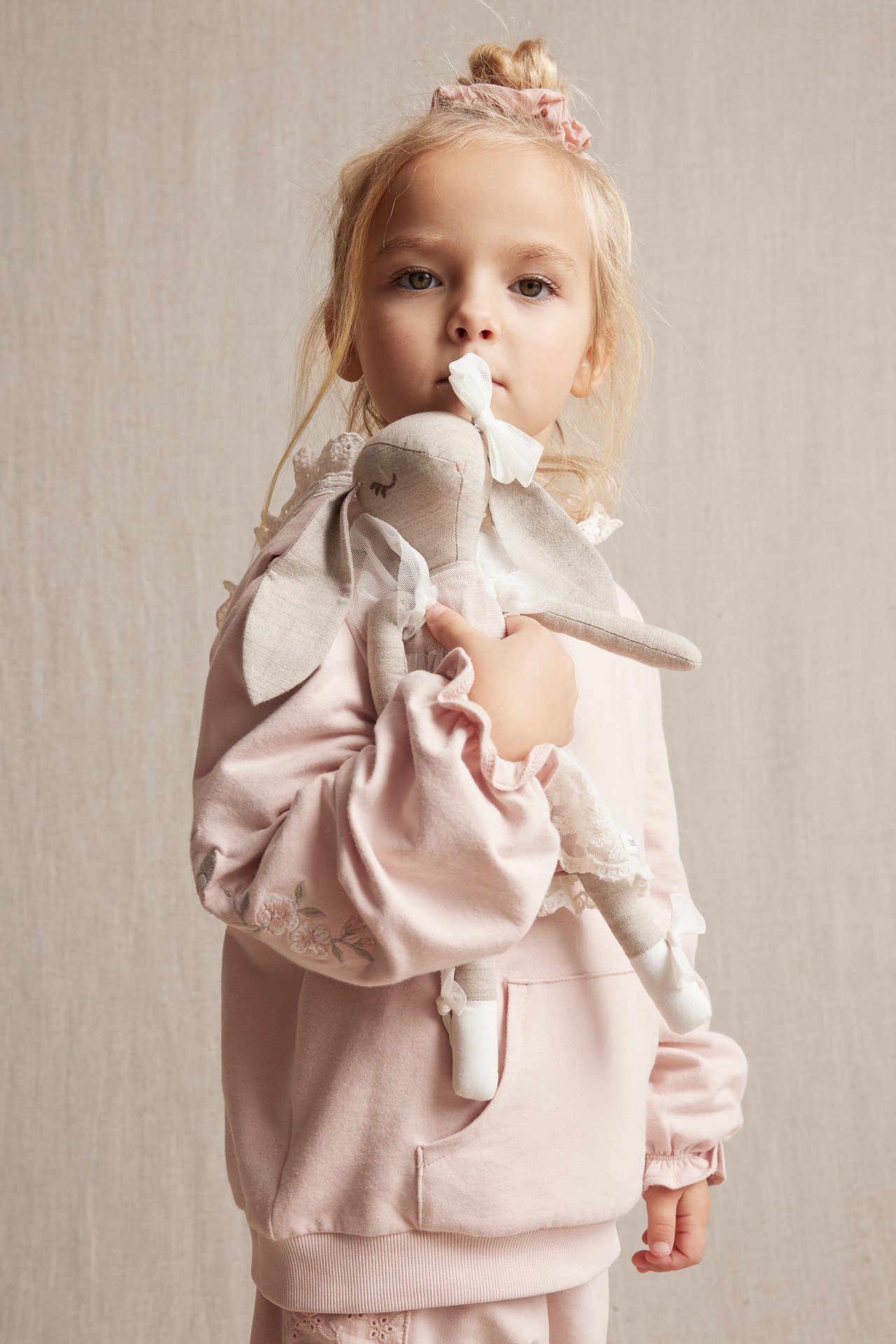 Front view of a kid in a light pink embroidered cotton hoodie, holding a bunny doll, with a pink scrunchie.