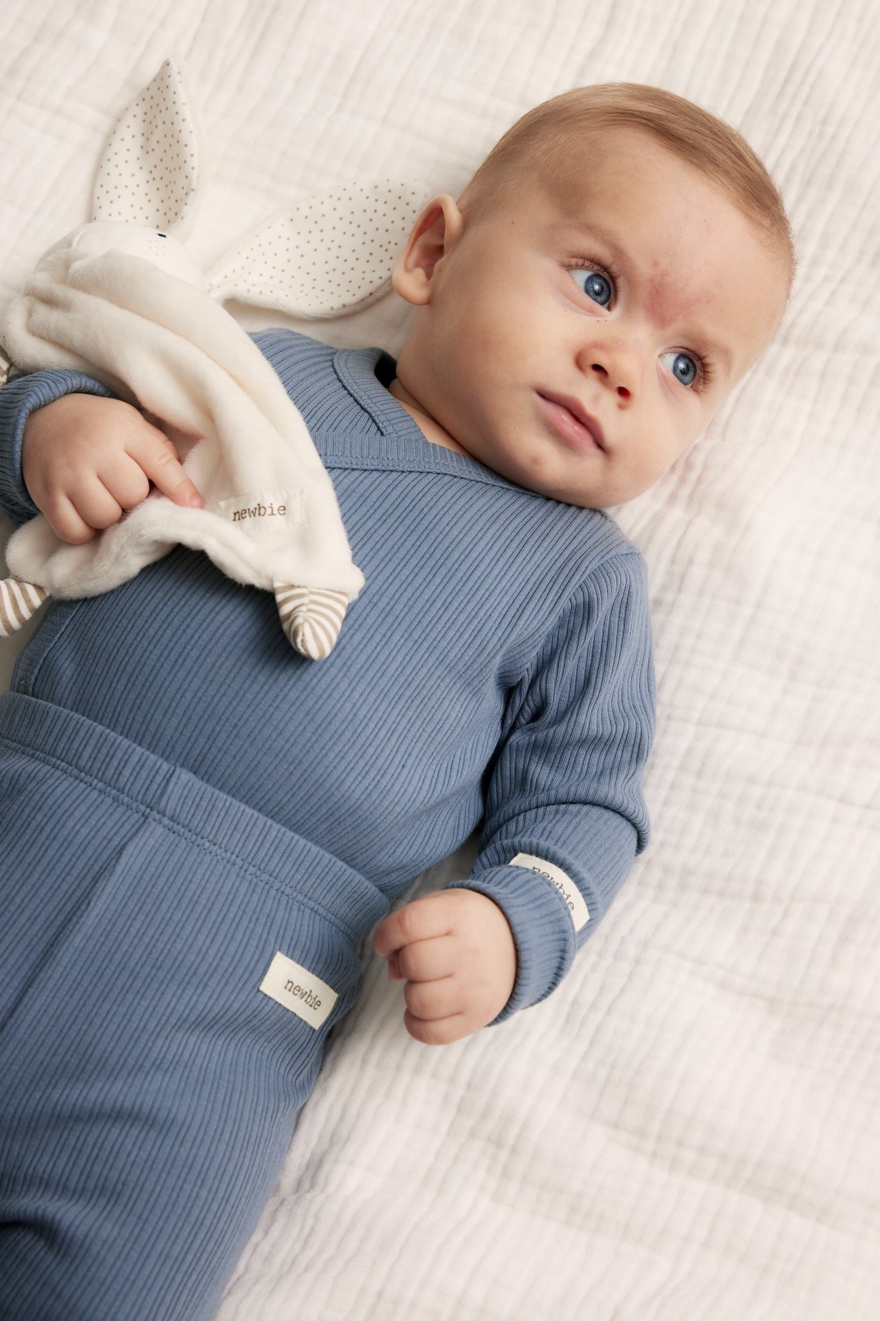 Close-up of a baby wearing a blue ribbed wrap bodysuit and matching pants, holding a white bunny comforter.