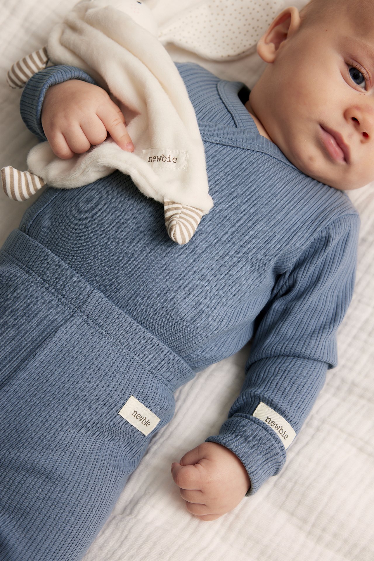 Close-up of a baby wearing a blue ribbed wrap bodysuit and matching pants, holding a white comfort blanket.