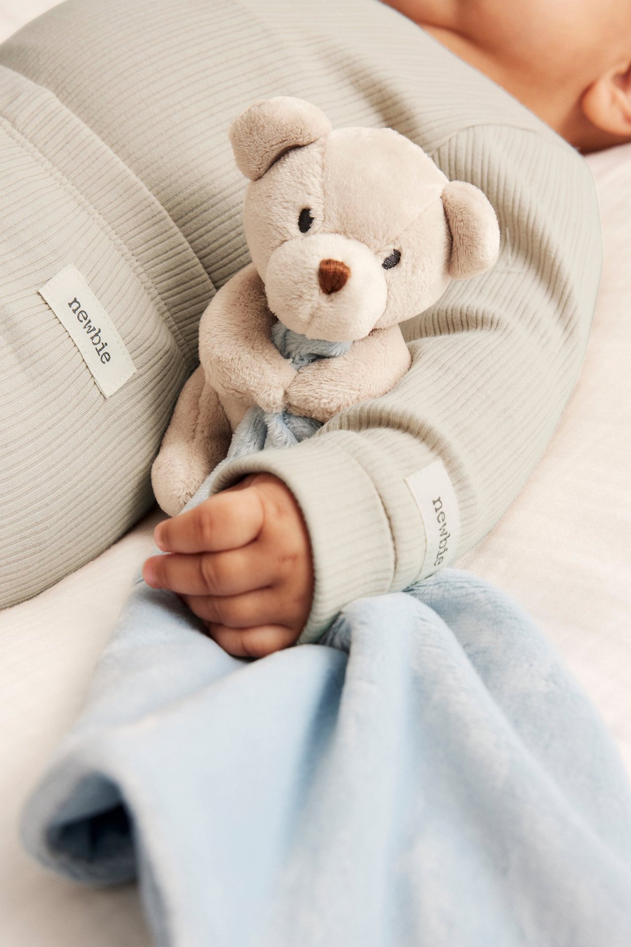 Nahaufnahme von Baby in hellgrünem Ripp-Body, hält beigen Teddybär und hellblaue Decke.