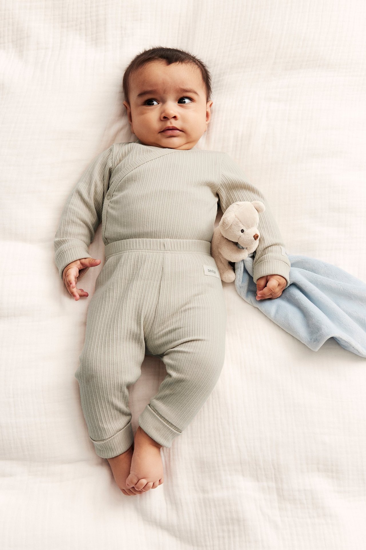 Draufsicht: Baby in beigem geripptem Wickelbody, passender Hose, hält Teddybär und blaue Decke.