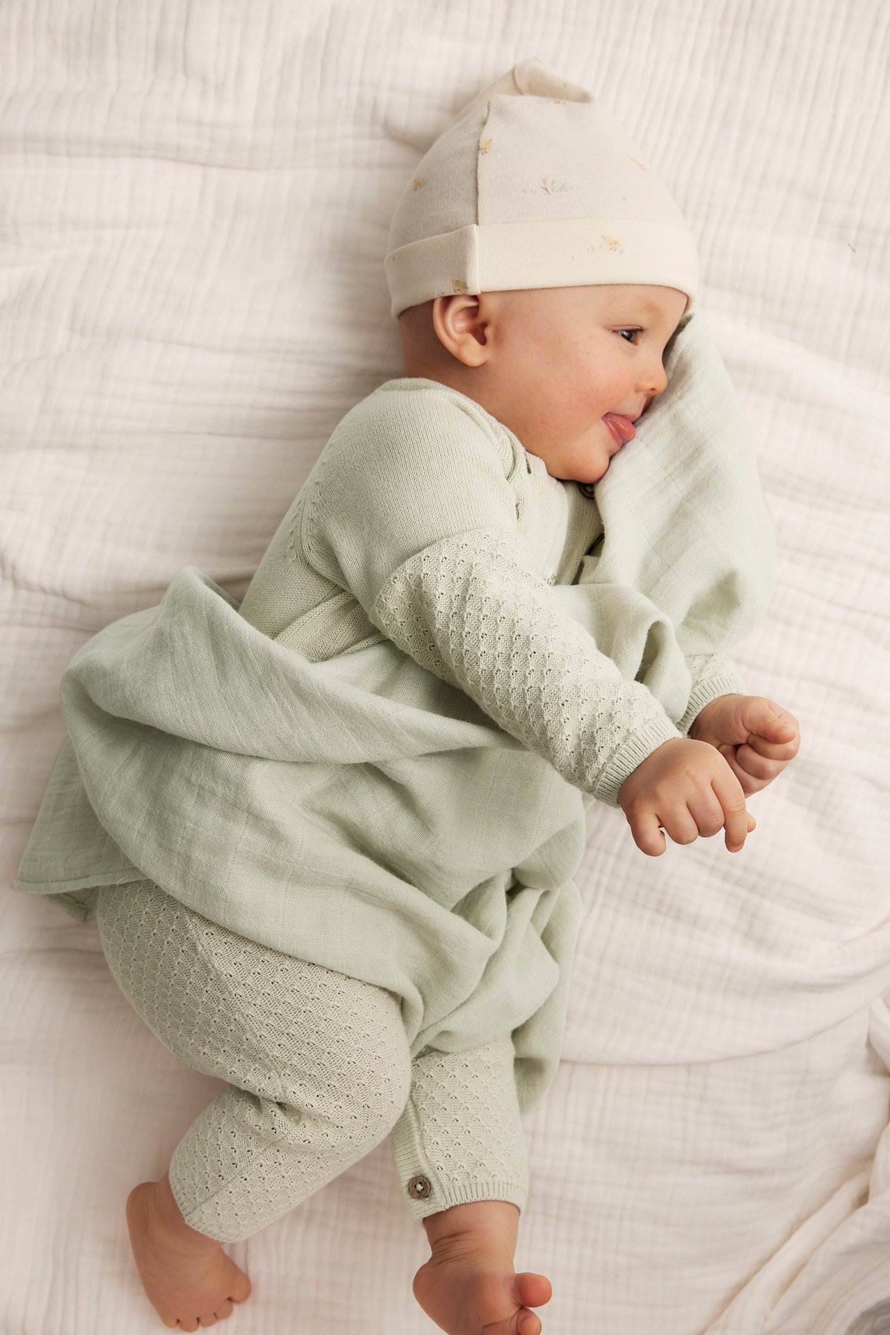 Top view: baby in light green textured knit jumpsuit, beige hat, and green blanket.