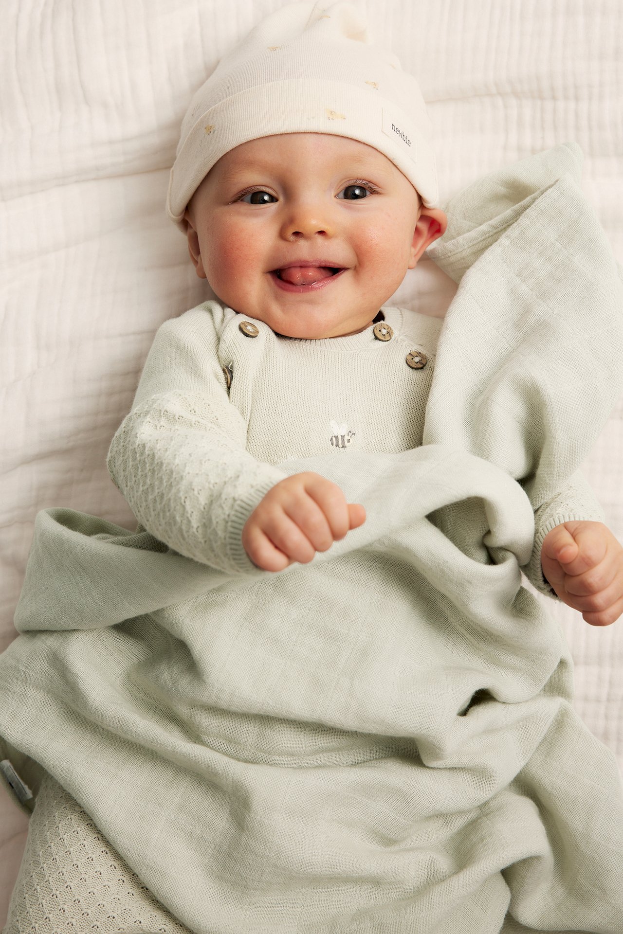 Smiling baby in a light green knitted cotton jumpsuit with buttons, embroidery, and a cream hat.