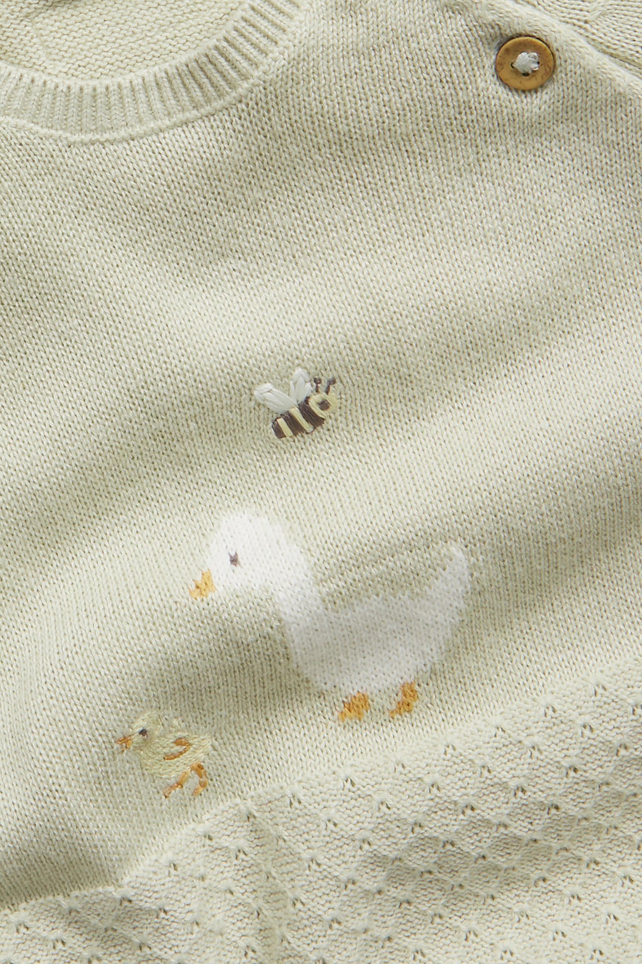 Close-up of a light green knitted baby jumpsuit with duck, chick, and bee embroidery, featuring a wooden button.