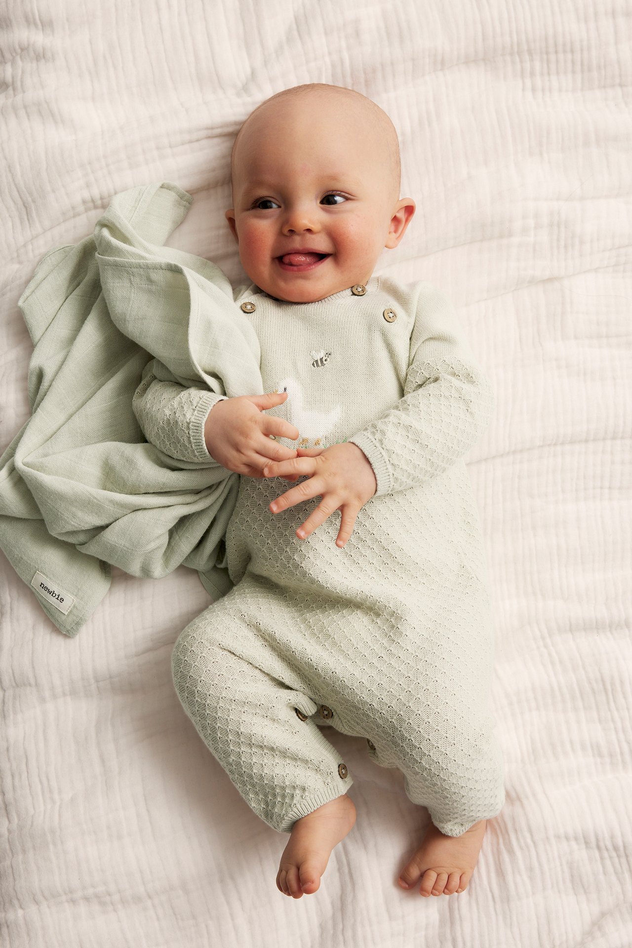 Top-down view of a smiling baby in a light green knitted jumpsuit with duck embroidery, holding a matching blanket.