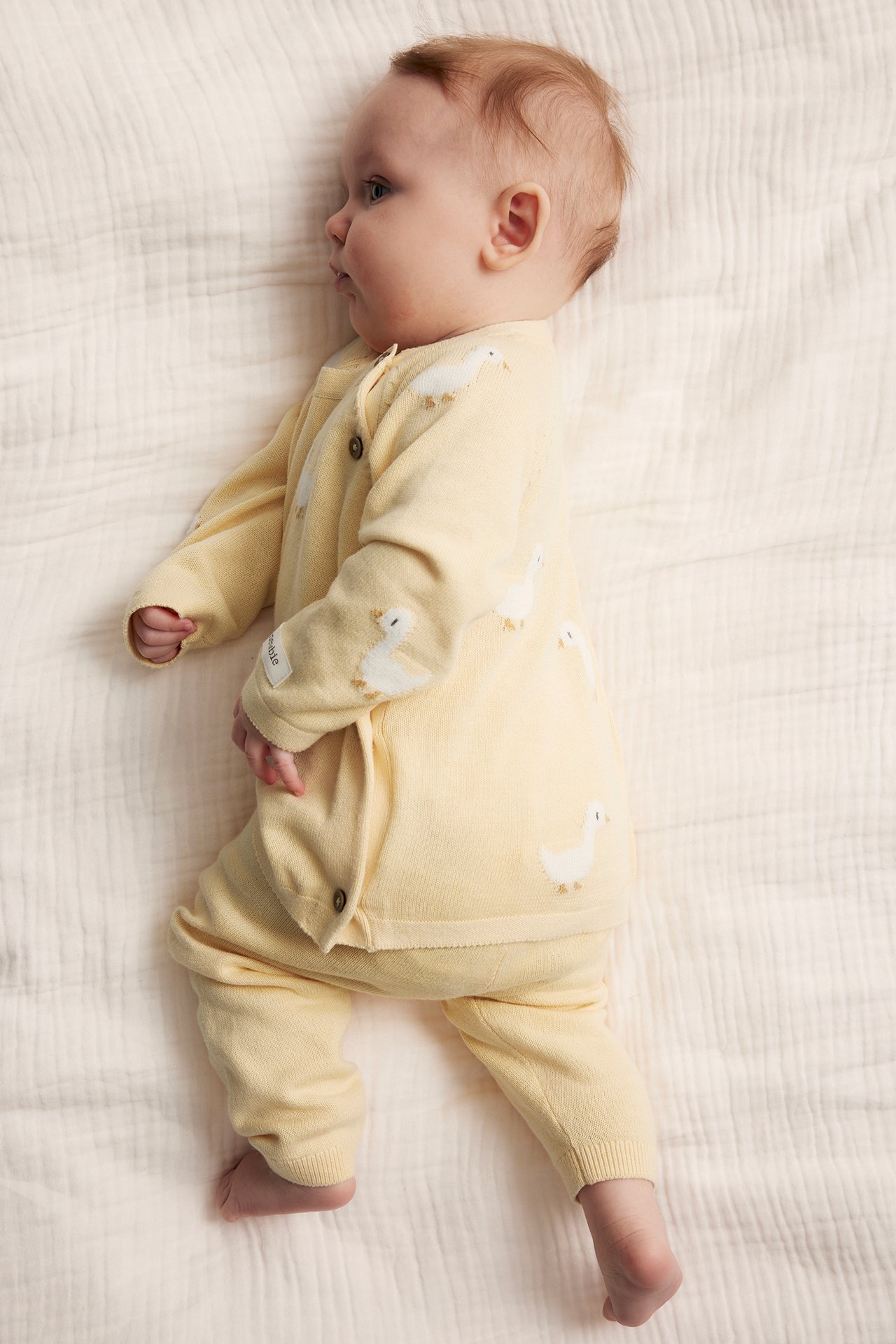 Side view of a baby in yellow knitted leggings and a matching yellow knitted cardigan with white duck patterns.