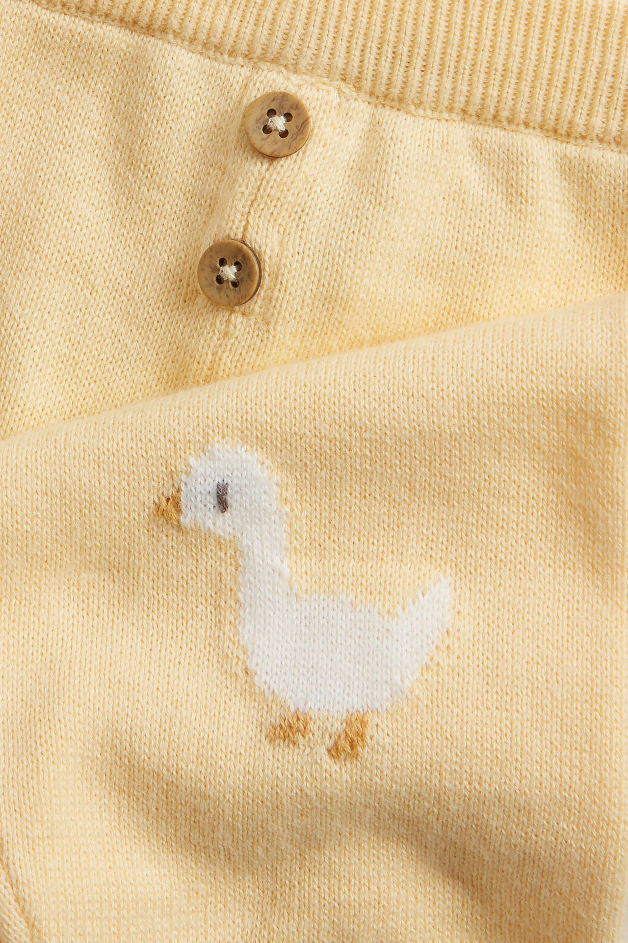 Nahaufnahme gelber gestrickter Baby-Leggings mit weißer Entenstickerei und zwei dekorativen Holzknöpfen.