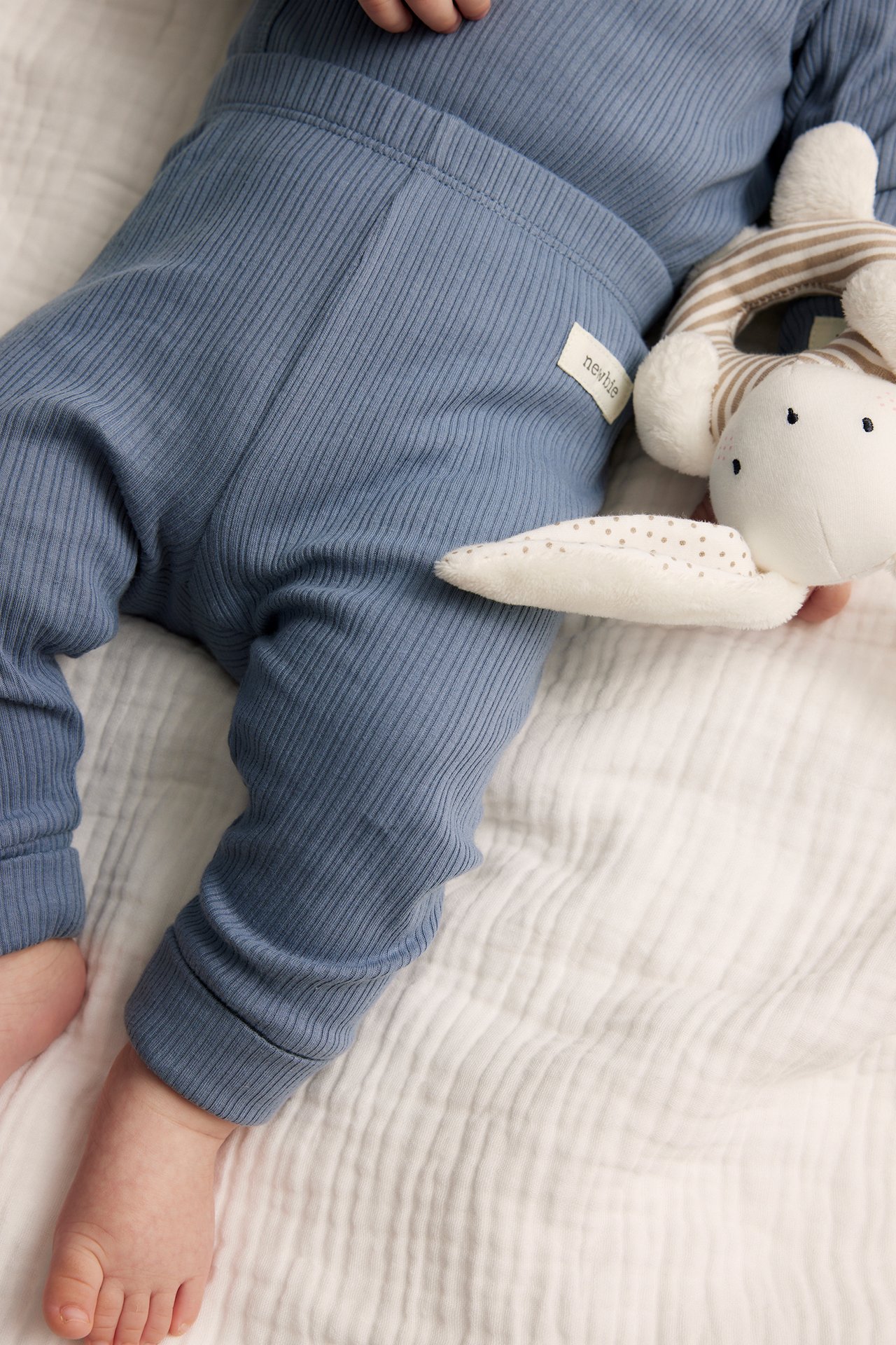 Close-up of a baby in blue ribbed leggings with folded cuffs, holding a striped stuffed animal.